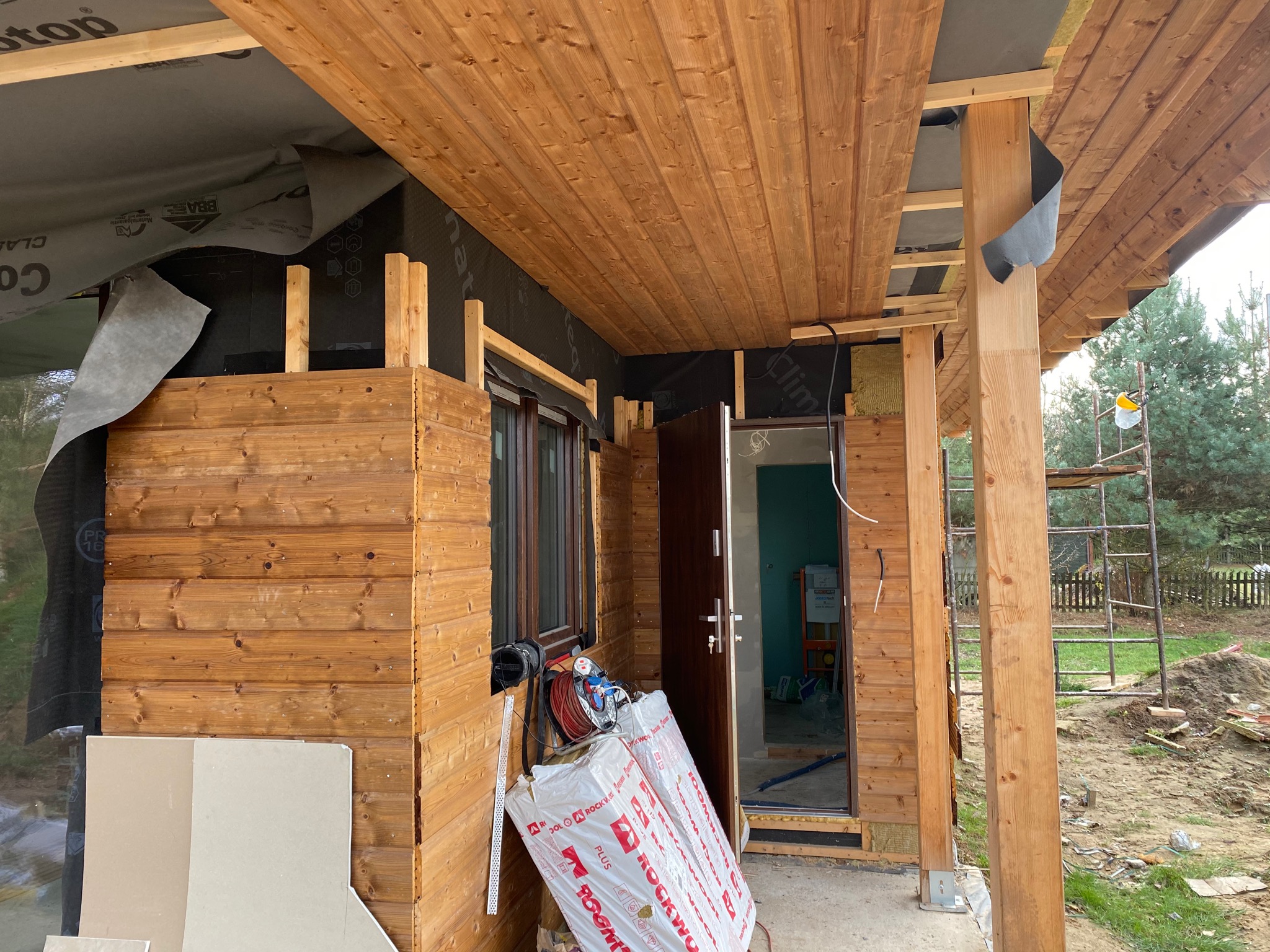 Wnętrze wiaty lub altany w trakcie budowy: widoczne drewniane deski elewacyjne, izolacja, folia paroizolacyjna, okno, drzwi wejściowe, rusztowanie w tle oraz materiały budowlane leżące na ziemi.