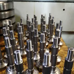 Partie metalowych, precyzyjnie obrobionych elementów z gwintem i okrągłą podstawą, ułożonych na drewnianej powierzchni, w tle widoczne metalowe pierścienie