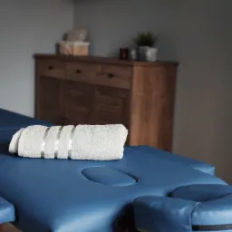 Niebieski stół do zabiegów z zagłówkiem i podłokietnikami, przykryty zwiniętym białym ręcznikiem z trzema srebrnymi paskami, w tle drewniana komoda i dekoracje.