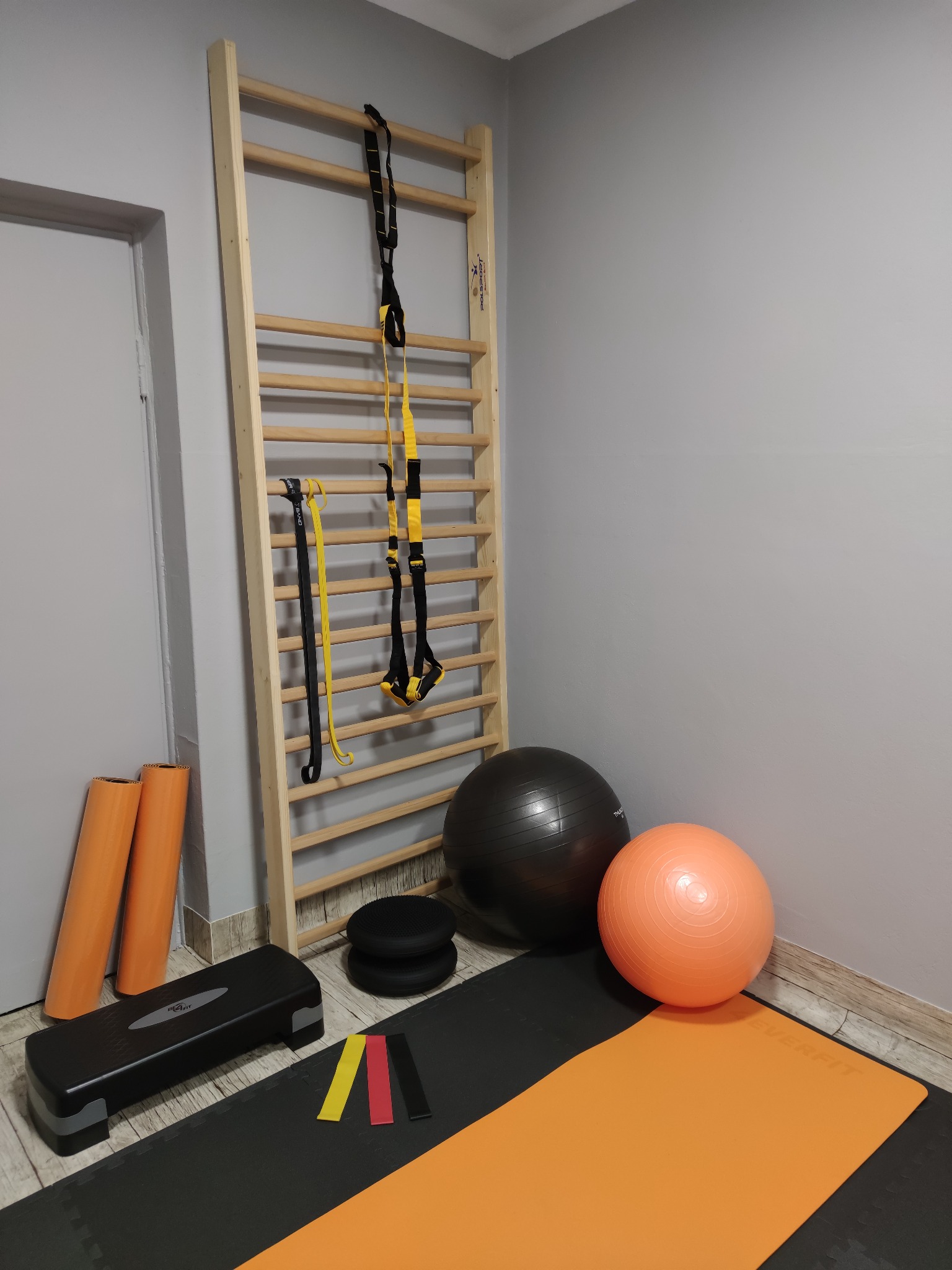 Ściana z drabinkami gimnastycznymi, piłkami do ćwiczeń, stepem i matą, przygotowana do zajęć rehabilitacyjnych.