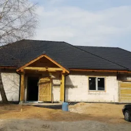 Surowy stan domu z białego pustaka z drewnianą konstrukcją wejścia i stertą materiałów budowlanych przed oknem, czarna dachówka i samochód na podjeździe.