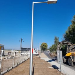 PM INSTALACJE Marcin Zimoch - Nowo zamontowana srebrna latarnia uliczna LED na tle błękitnego nieba, widoczny wykop, ogrodzenie z siatki, pracująca koparka i ekipa robotników w oddali.