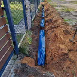 PM INSTALACJE Marcin Zimoch - Wykopany rów wyłożony niebieską folią ochronną wzdłuż ogrodzenia posesji, przygotowany do instalacji podziemnej, z widoczną łopatą i pracownikami w oddali.