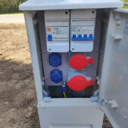 Otwarta skrzynka elektryczna z bezpiecznikami, gniazdami siłowymi (czerwone) i standardowymi (niebieskie), zamontowana na zewnątrz, widoczne przewody. Skrzynka umieszczona na ziemi.