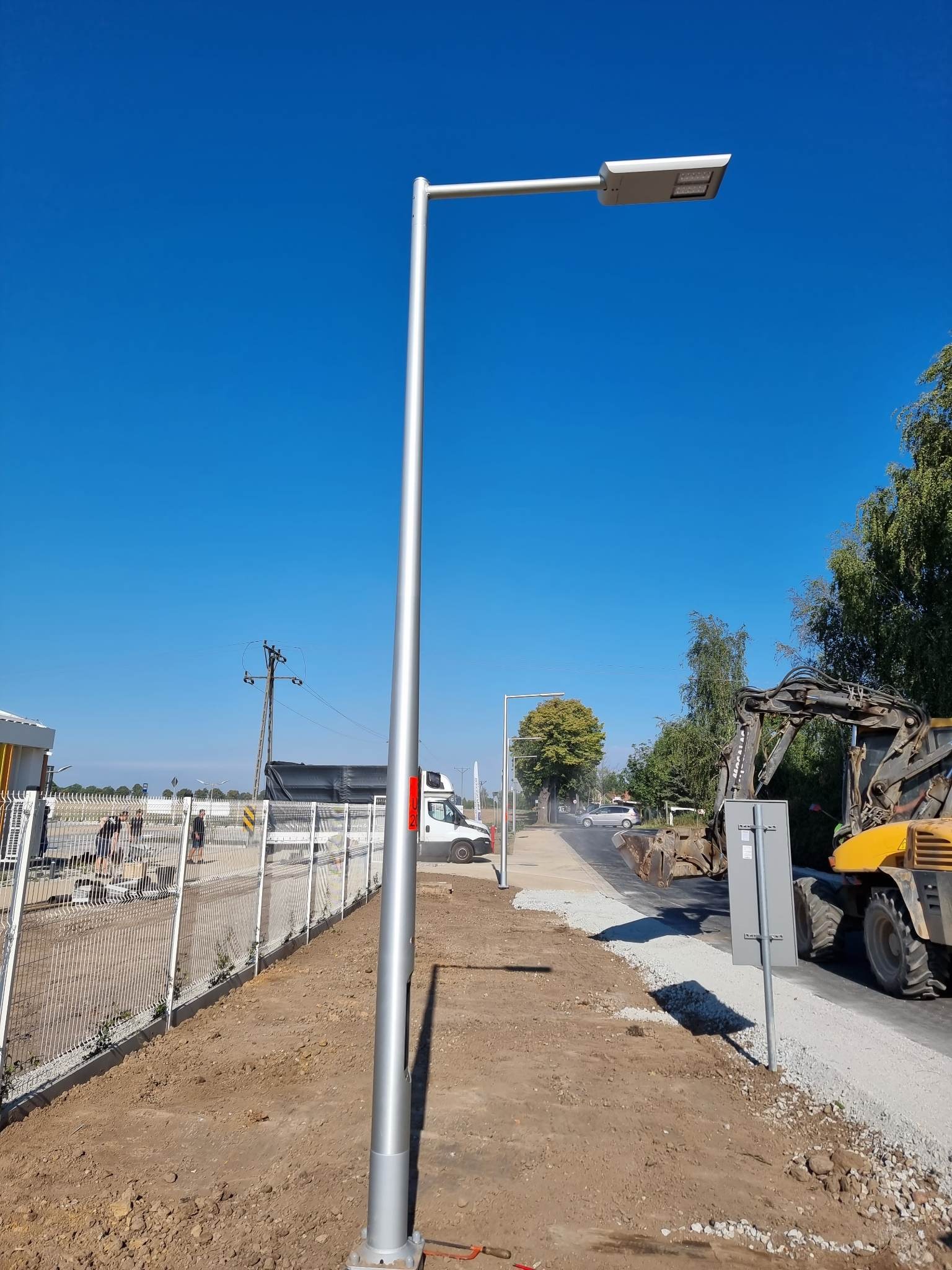 Nowo zamontowana srebrna latarnia uliczna LED na tle błękitnego nieba, widoczny wykop, ogrodzenie z siatki, pracująca koparka i ekipa robotników w oddali.