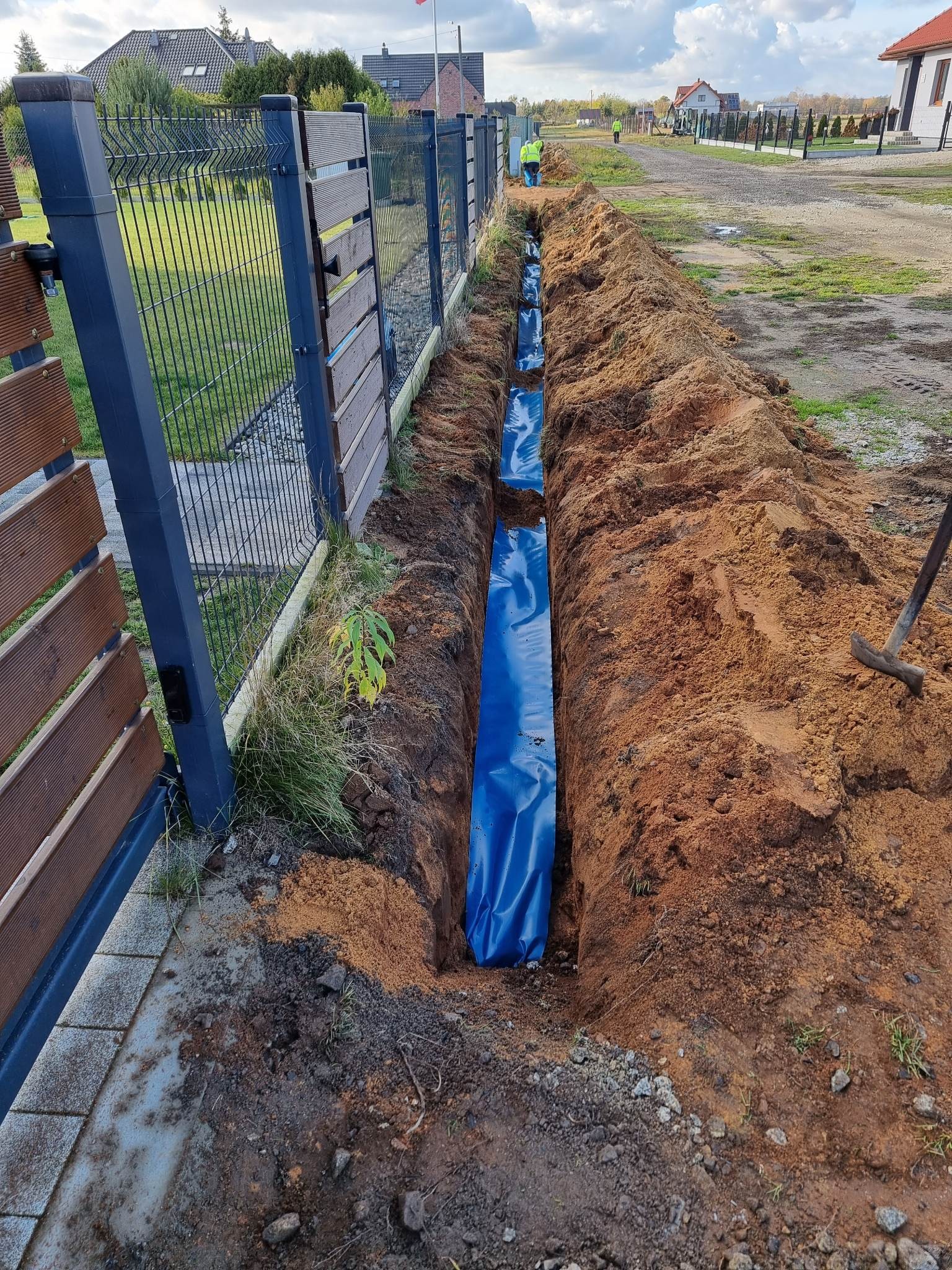 Wykopany rów wyłożony niebieską folią ochronną wzdłuż ogrodzenia posesji, przygotowany do instalacji podziemnej, z widoczną łopatą i pracownikami w oddali.