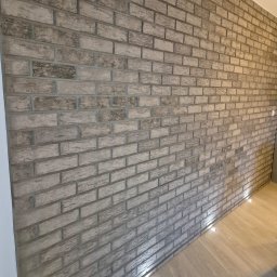 LEVEL-BUD WOJCIECH JANIK - Ściana wykończona dekoracyjną cegłą w odcieniach szarości, z subtelnym oświetleniem podłogowym. Widok pod kątem, ukazujący strukturę i fakturę materiału.