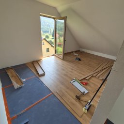 LEVEL-BUD WOJCIECH JANIK - Poddasze w trakcie układania paneli podłogowych. Widoczne narzędzia, podkład i częściowo ułożone panele. W tle okno z widokiem na zielone wzgórza i zabudowania.