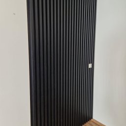 LEVEL-BUD WOJCIECH JANIK - Ciemne lamele na ścianie, z widocznymi gniazdkami elektrycznymi i fragmentem podłogi z paneli, tworzące nowoczesny element dekoracyjny wnętrza.