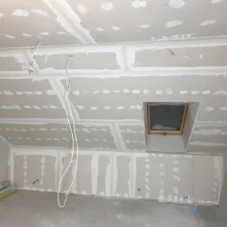 Poddasze w trakcie wykańczania: zamontowane płyty gipsowo-kartonowe, widoczne łączenia i szpachlowanie, okno dachowe, przewody elektryczne zwisające z sufitu.