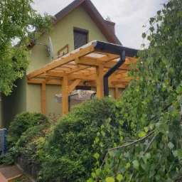 Drewniana konstrukcja z przezroczystym zadaszeniem, prawdopodobnie z poliwęglanu, przylegająca do ściany domu w kolorze oliwkowym, z widoczną rynną, otoczona zielenią.