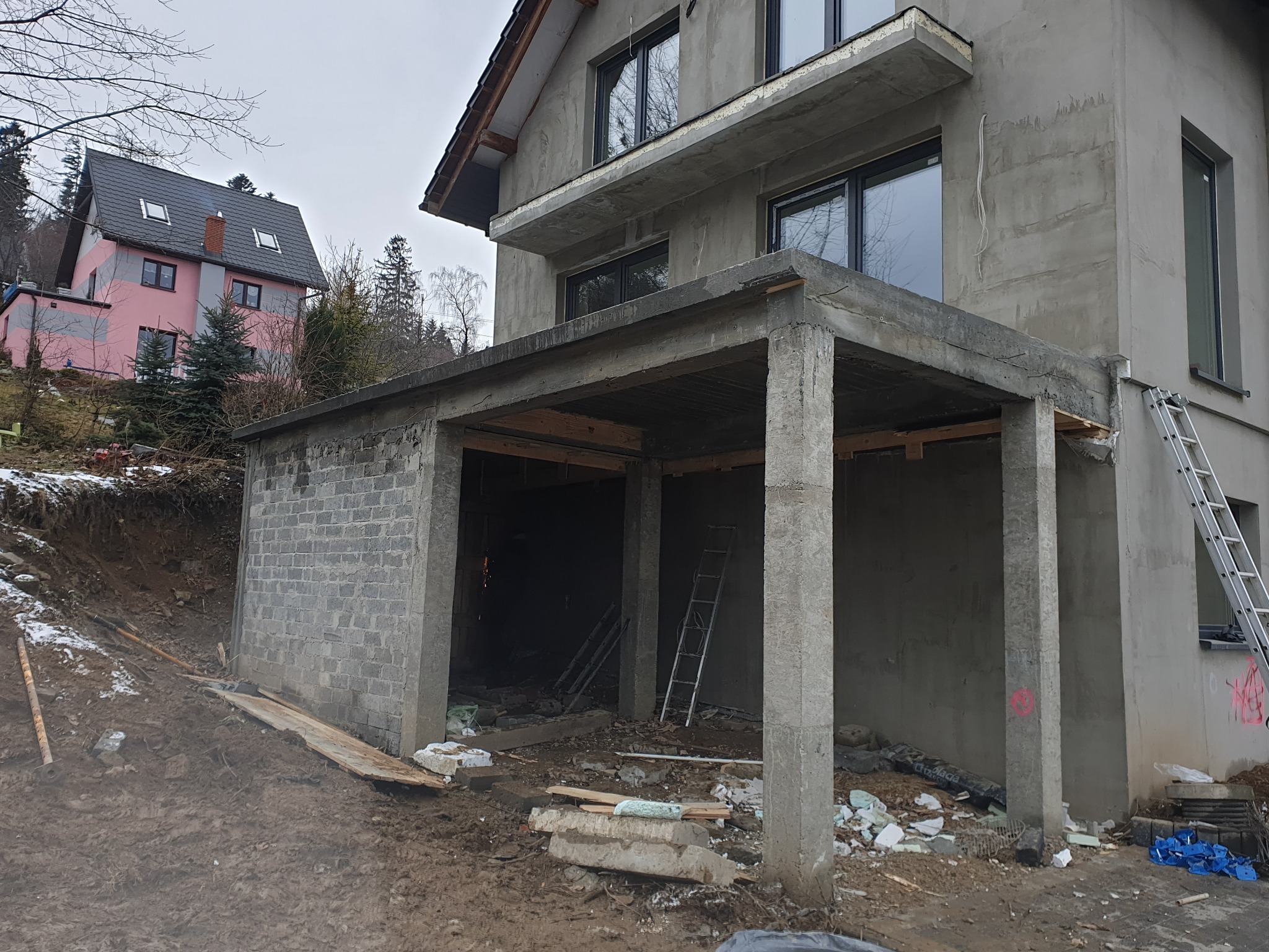 Surowy stan budynku w trakcie budowy z widocznym zadaszeniem tarasu, nieotynkowane ściany, gruz i drabina oparta o ścianę.