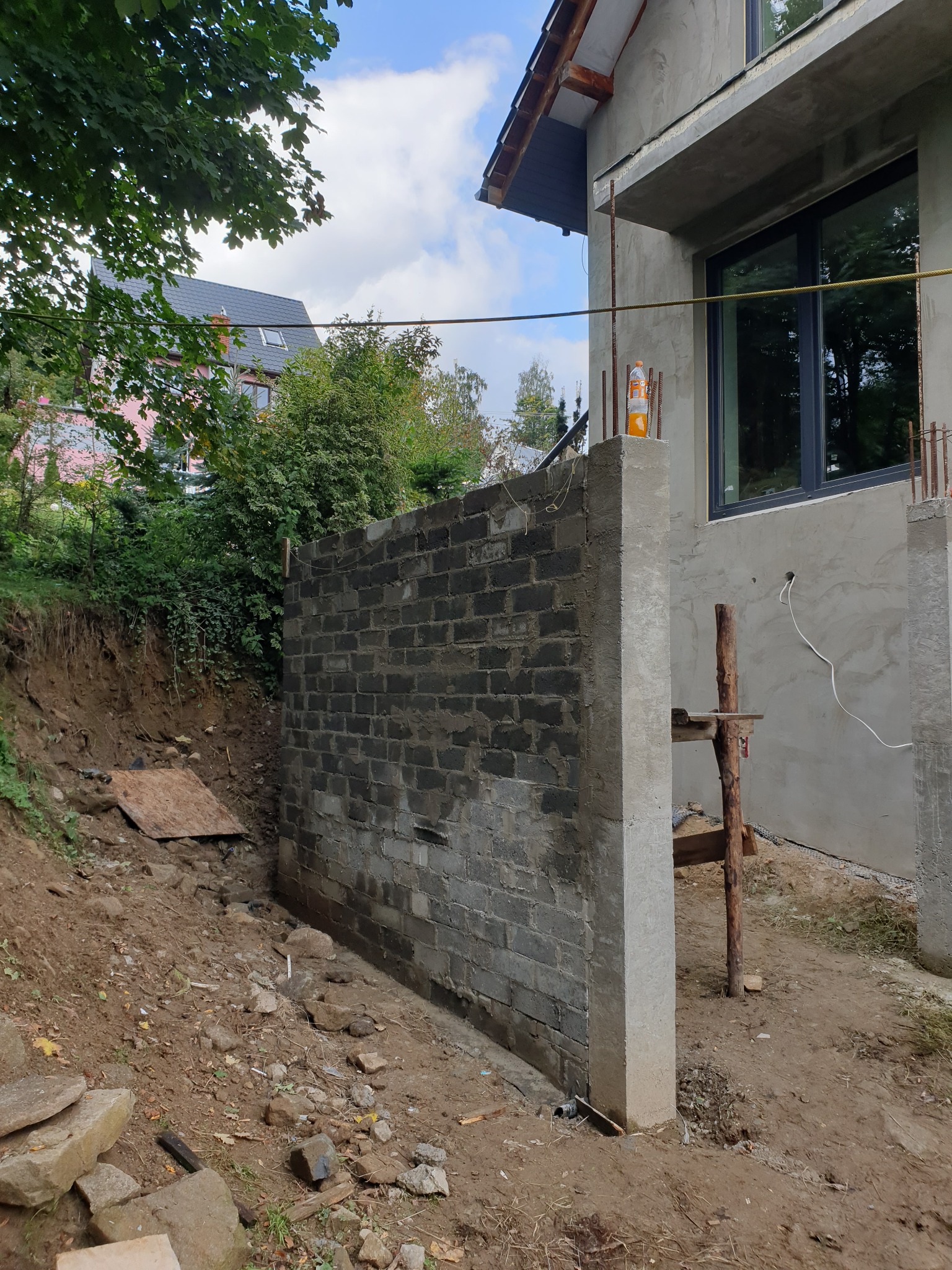 Wznoszenie muru oporowego z betonowych bloczków przy budynku w trakcie budowy, widoczna ziemia i prowizoryczne rusztowanie.