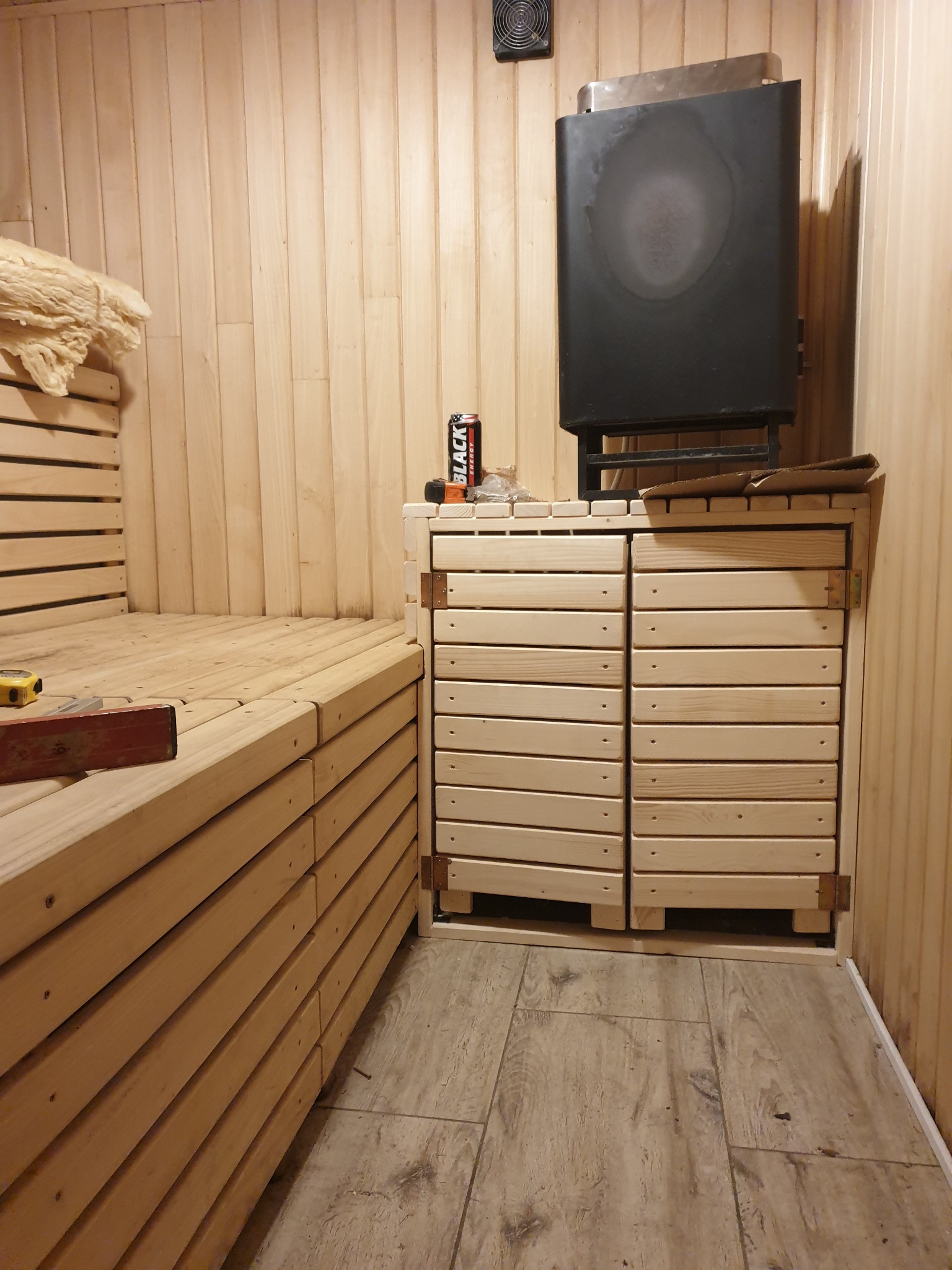 Wnętrze sauny w trakcie budowy, widoczne drewniane ławy, piec, obudowa pieca z drewnianymi drzwiczkami, poziomica i miarka na ławie, puszka napoju energetycznego na obudowie pieca.