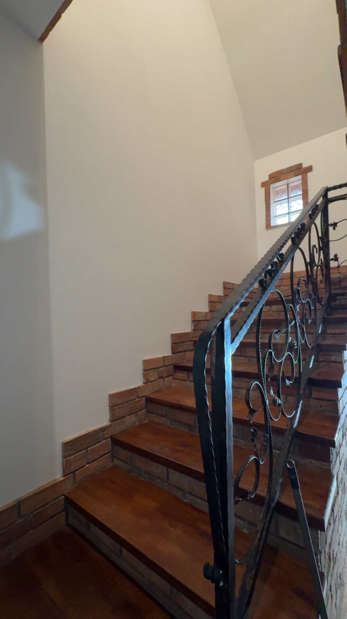 Schody z drewnianymi stopniami i ceglanym wykończeniem, ozdobna metalowa balustrada. Widok z dołu, okno w tle.