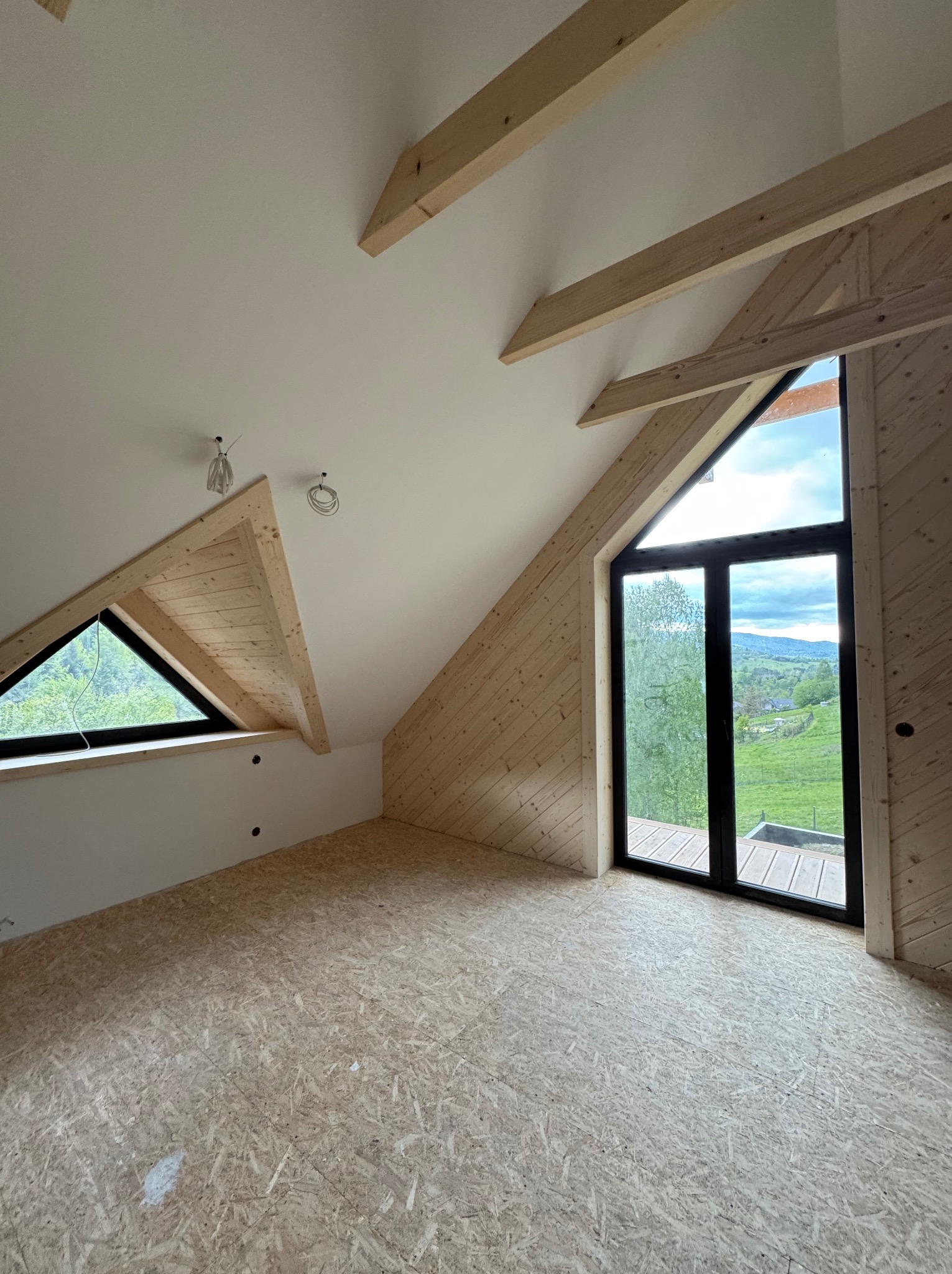 Poddasze z drewnianą boazerią, oknami trójkątnymi i podłogą z płyt OSB. Widoczne elementy konstrukcyjne dachu i instalacje elektryczne. W tle zielony krajobraz.