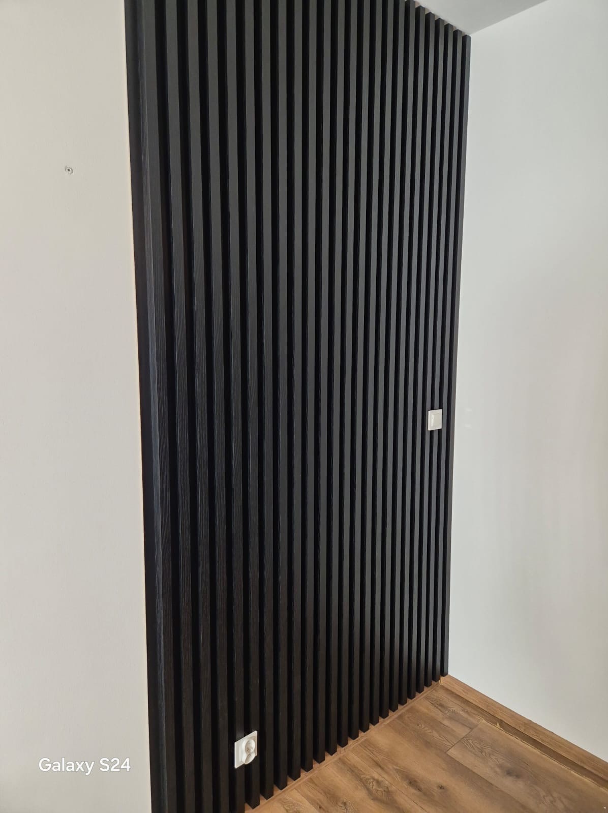 Ciemne lamele na ścianie, z widocznymi gniazdkami elektrycznymi i fragmentem podłogi z paneli, tworzące nowoczesny element dekoracyjny wnętrza.