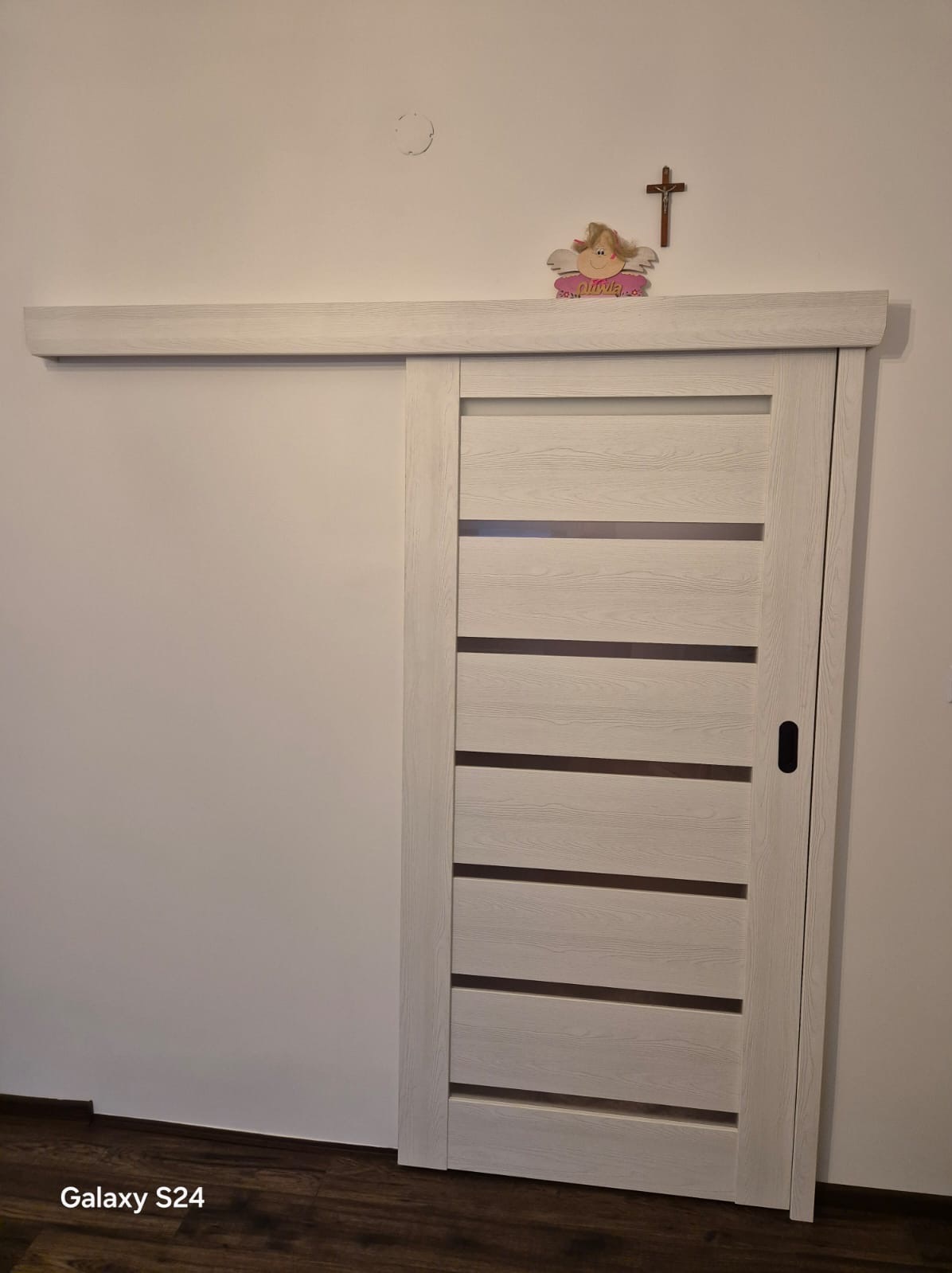 Białe, przesuwne drzwi z poziomymi przeszkleniami, zamontowane w ścianie. Nad drzwiami dekoracje: aniołek i krzyż. Widoczna podłoga z paneli.