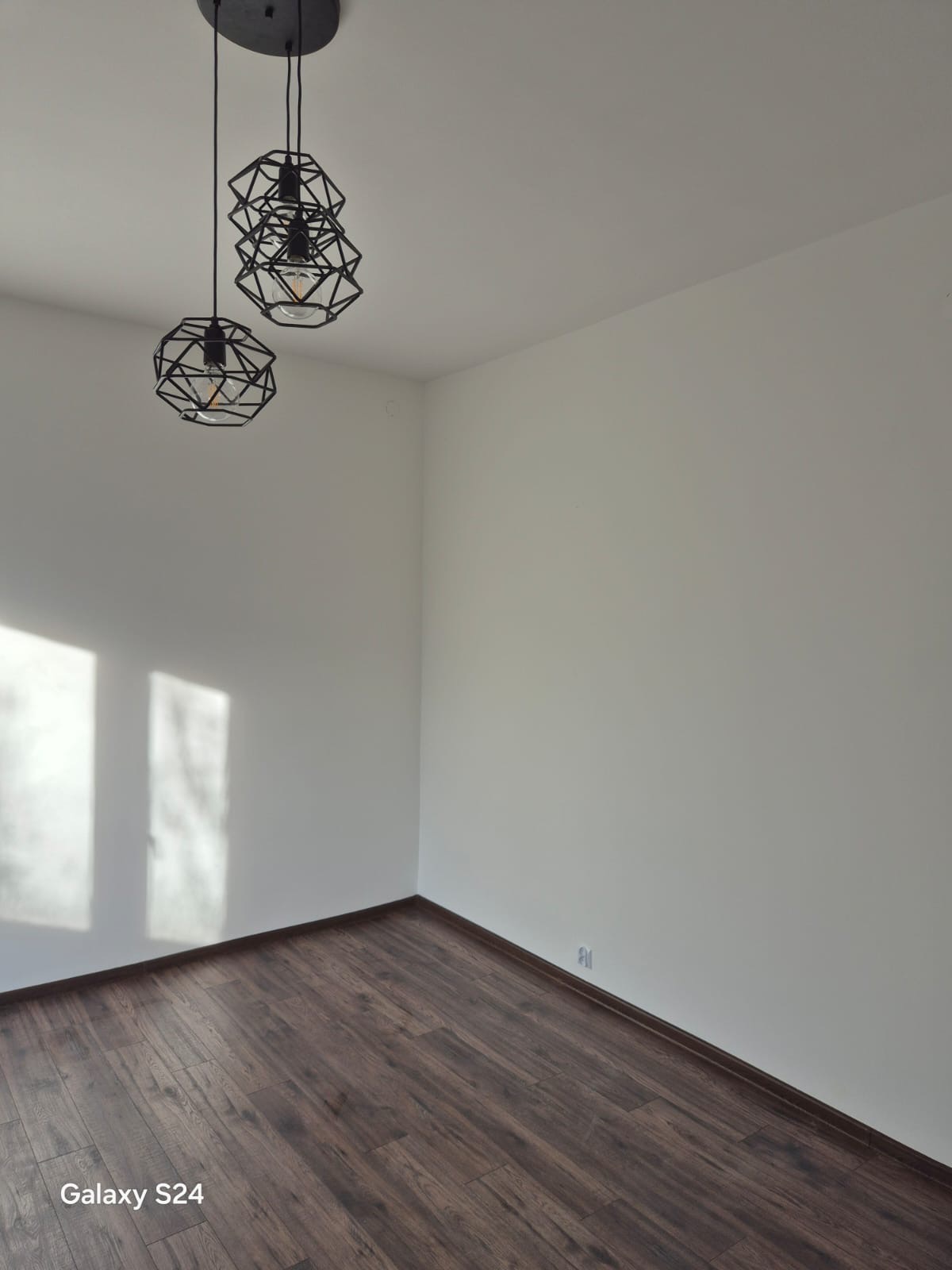 Wnętrze pokoju po remoncie: ciemne panele podłogowe, białe ściany i nowoczesne, geometryczne lampy wiszące. Widoczne światło słoneczne wpadające przez okno.
