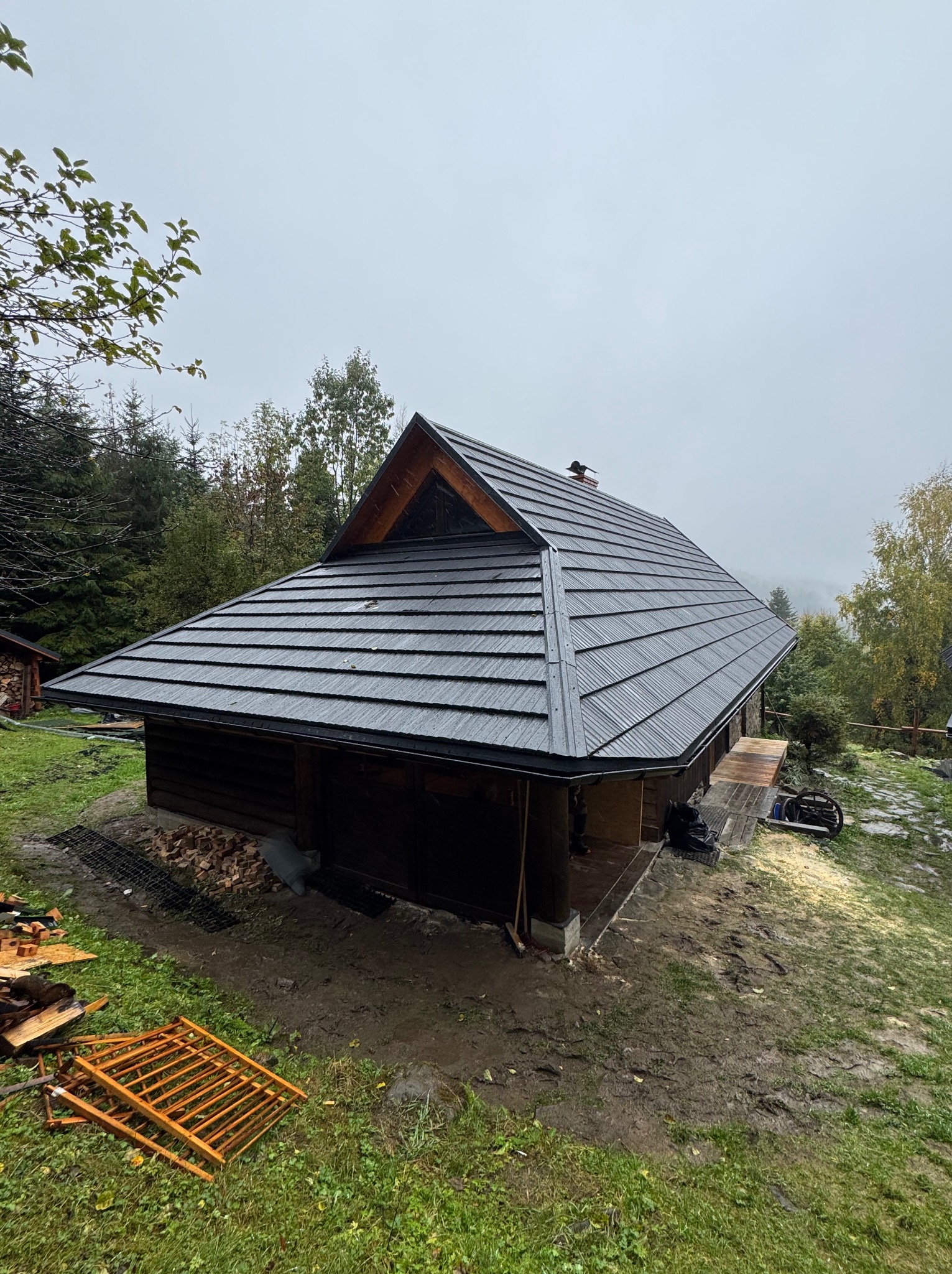 Drewniany dom z ciemną, nową dachówką na tle zalesionego wzgórza w pochmurny dzień. Widoczne elementy konstrukcyjne i otoczenie domu.