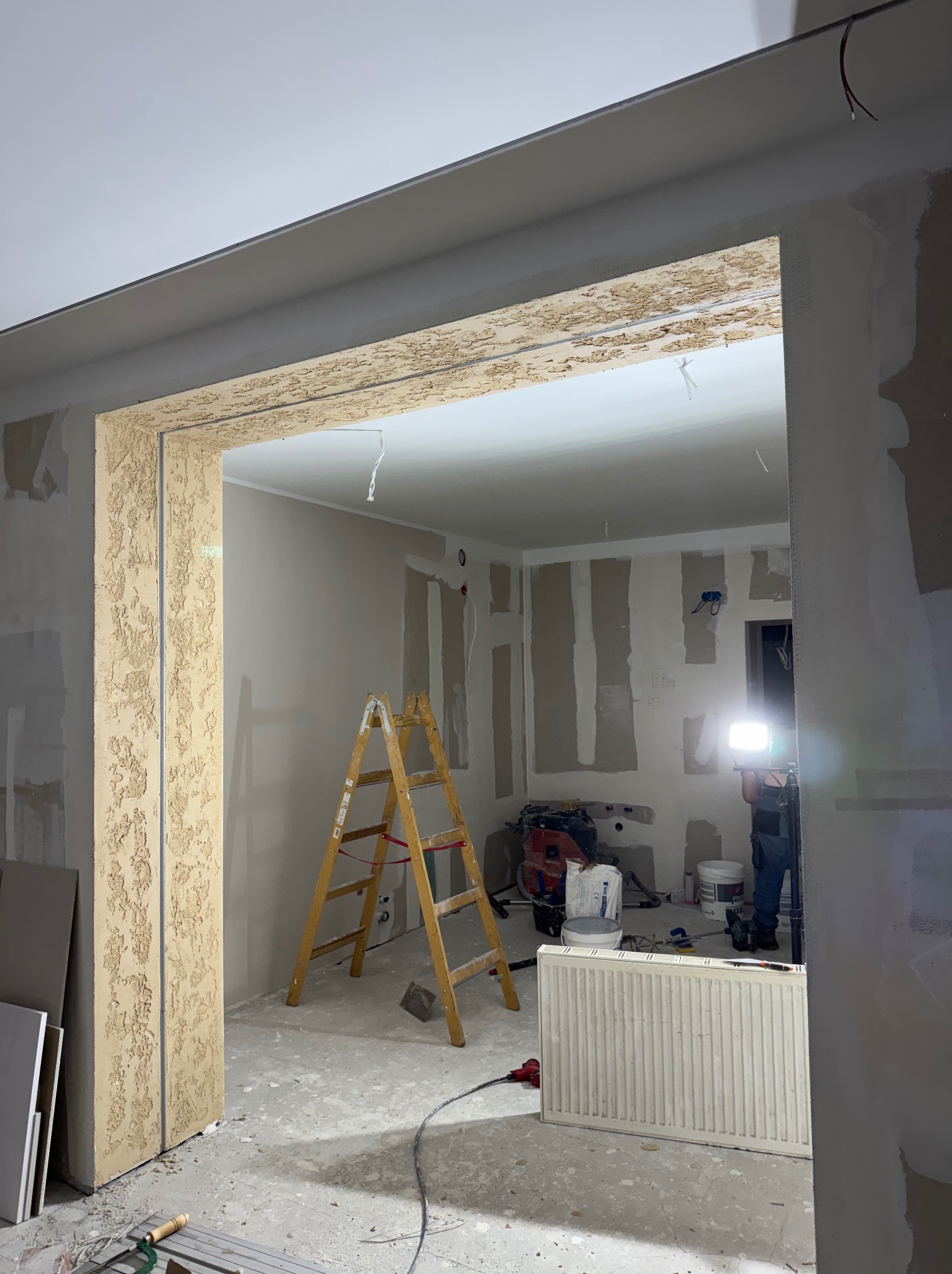 Wnętrze w trakcie remontu: ściany z płyt gipsowych, drabina, grzejnik, widoczne narzędzia i osoba z lampą oświetlająca przestrzeń. Fragment dekoracyjnej faktury na ścianie.