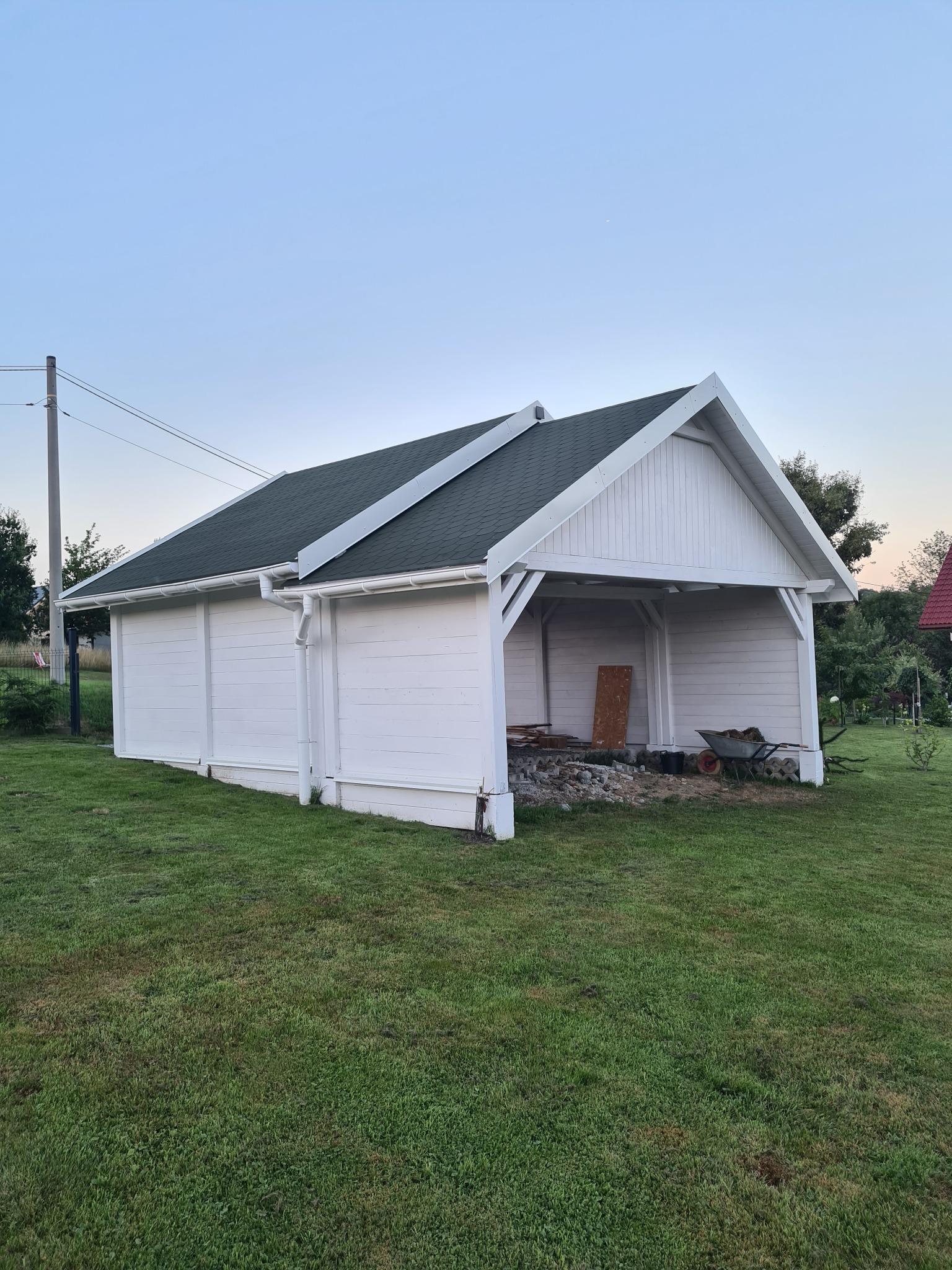 Biała, drewniana altana z zielonym dachem w trakcie budowy, widoczne materiały budowlane i taczka na trawniku.