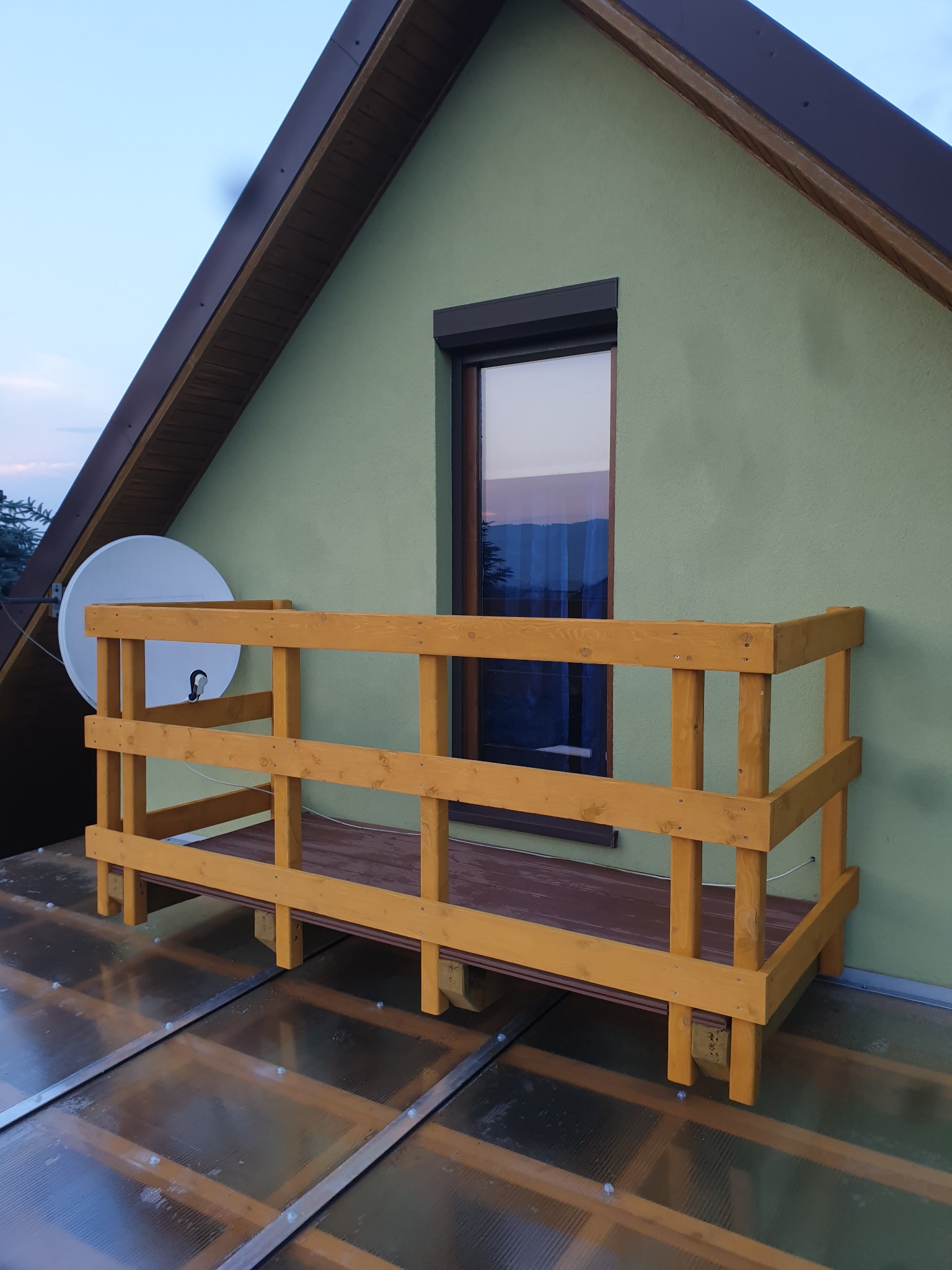 Nowy, drewniany balkon z żółtą balustradą, zamontowany na przezroczystym zadaszeniu tarasu przy zielonym domu z anteną satelitarną.