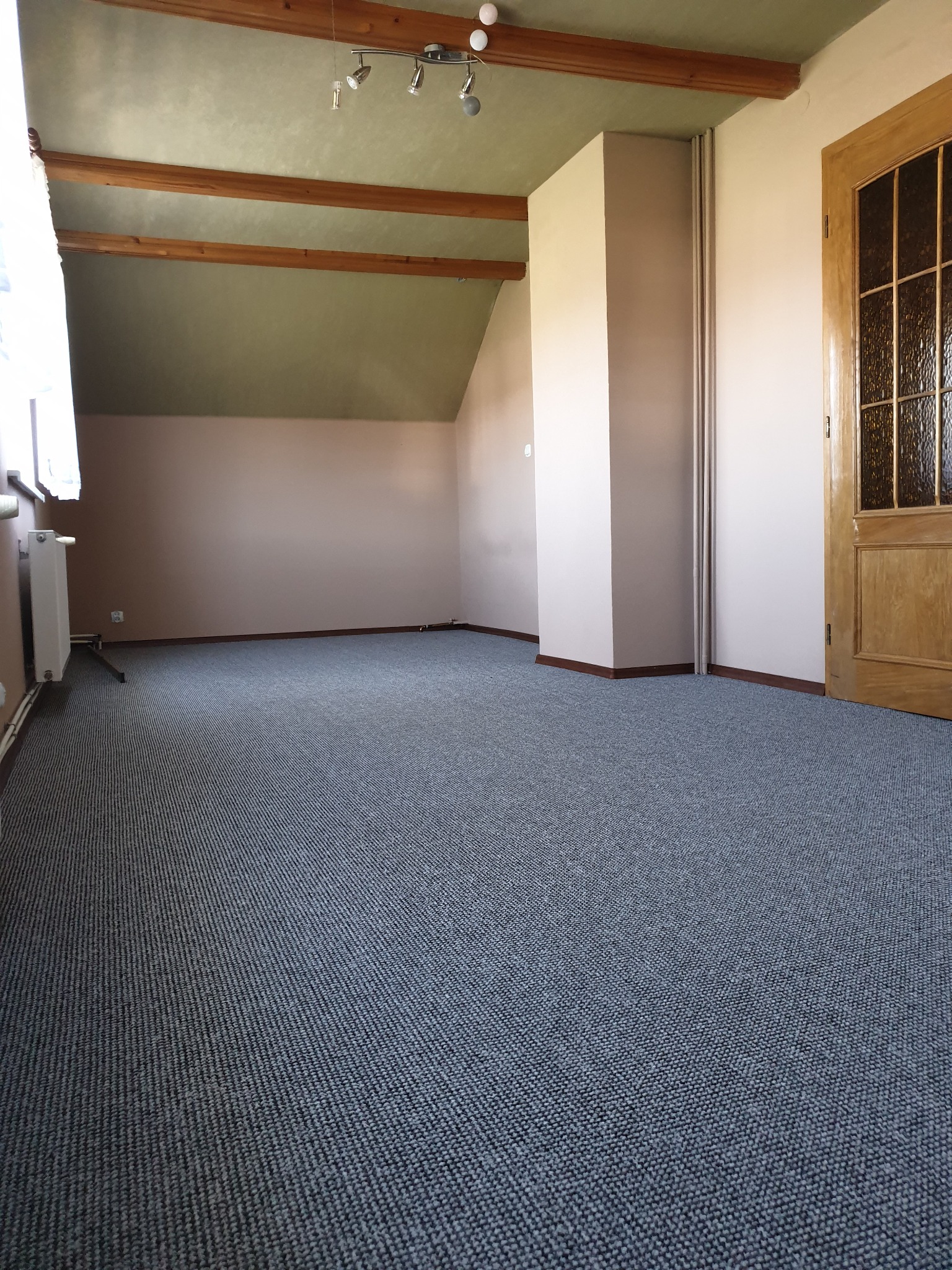 Pokój na poddaszu z nową, szarą wykładziną dywanową, drewnianymi belkami stropowymi i oknem po lewej stronie.