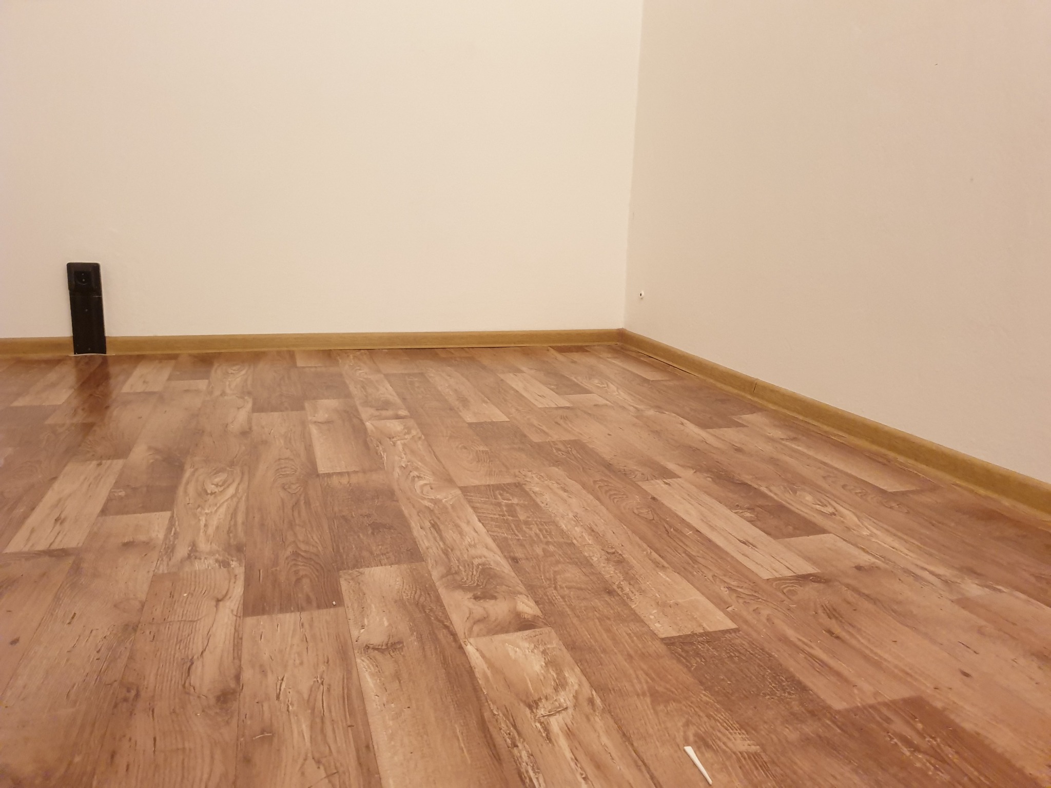 Ukończone układanie paneli podłogowych w rogu pokoju, widoczna listwa przypodłogowa i czarny ogranicznik do drzwi przy ścianie.