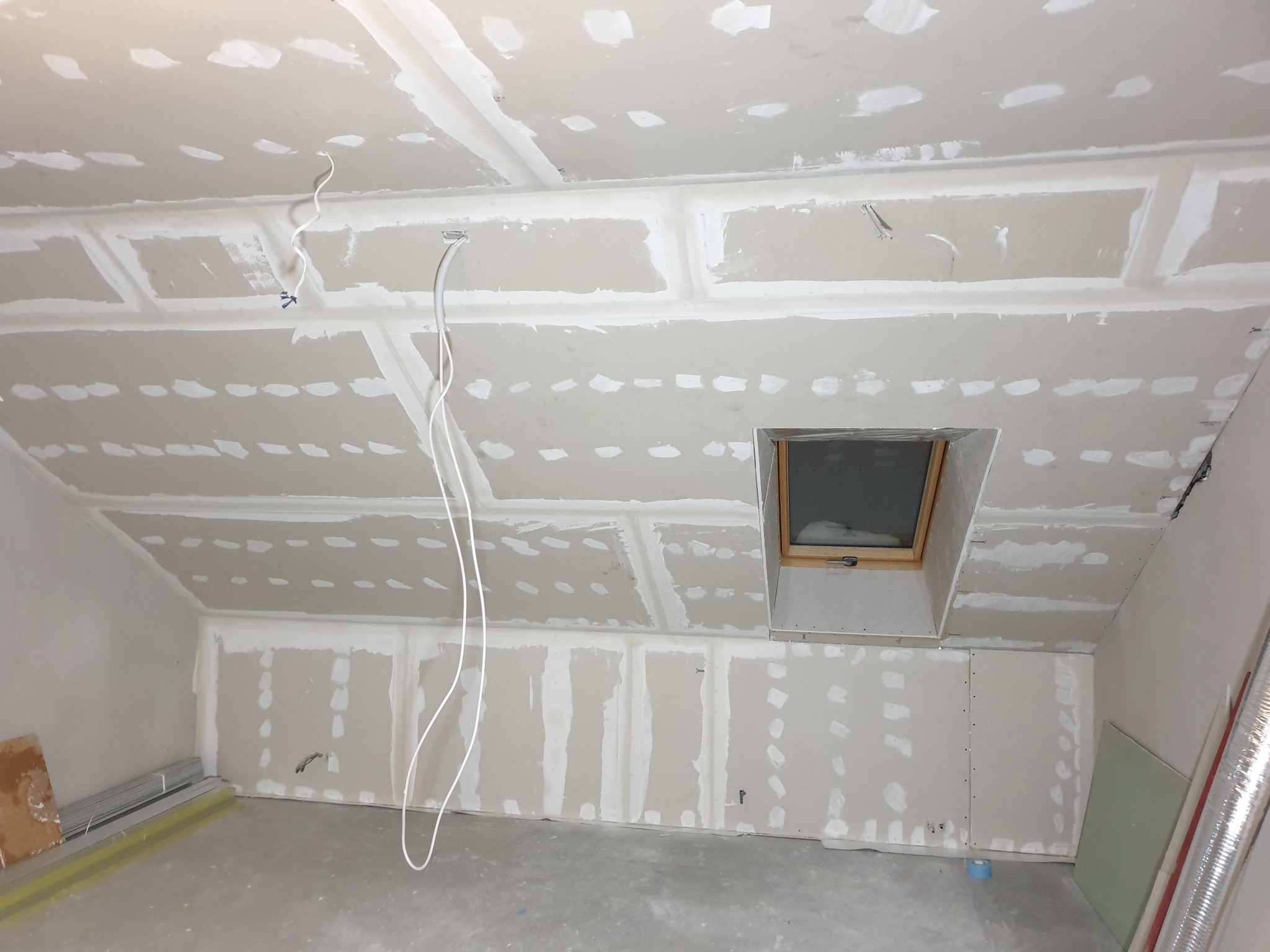 Poddasze w trakcie wykańczania: zamontowane płyty gipsowo-kartonowe, widoczne łączenia i szpachlowanie, okno dachowe, przewody elektryczne zwisające z sufitu.