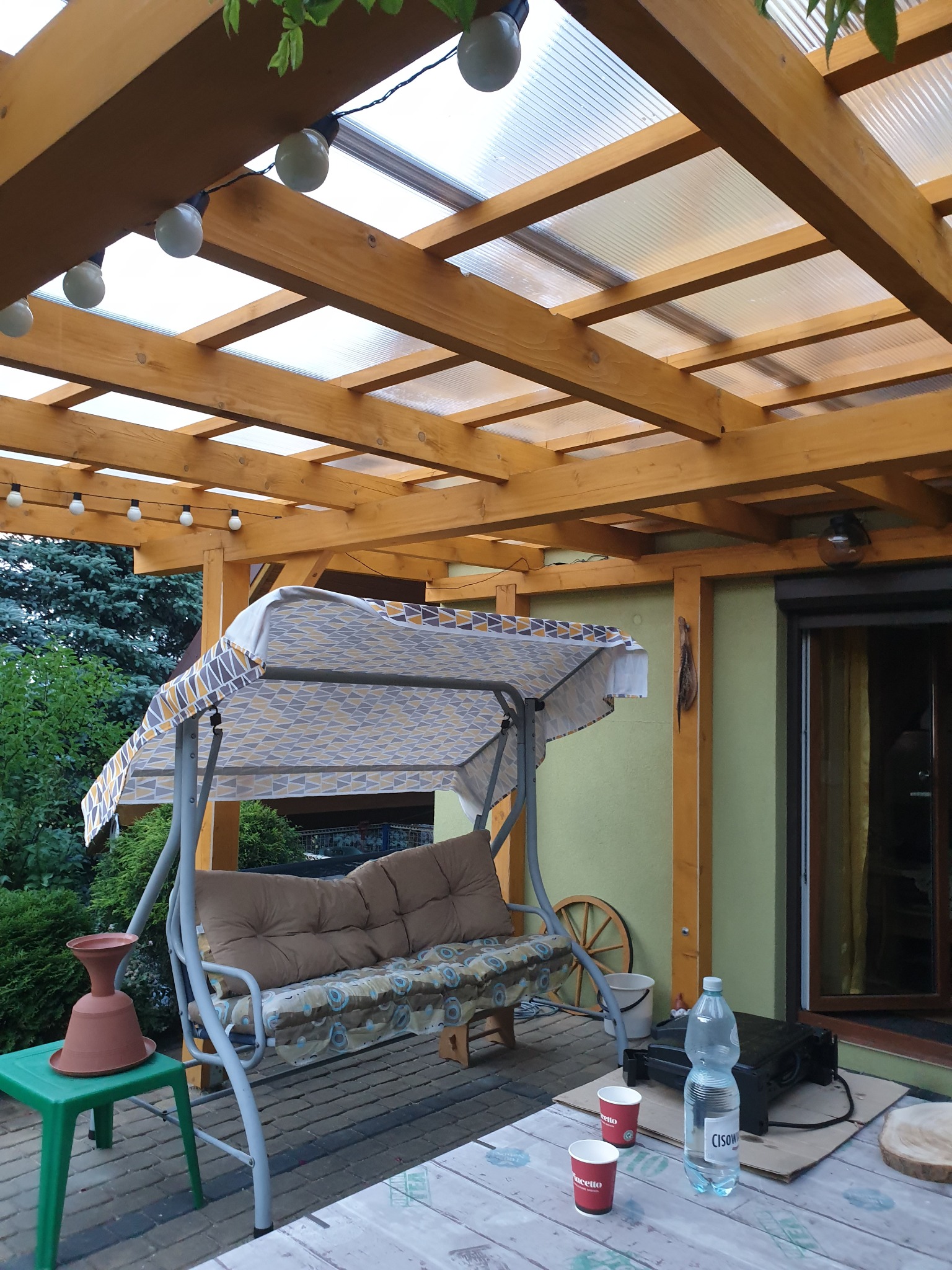 Zadaszony taras z drewnianą konstrukcją, oświetlony girlandą żarówek, z huśtawką ogrodową, stolikiem, grillem elektrycznym i dekoracjami, widok na zieleń ogrodu w tle.