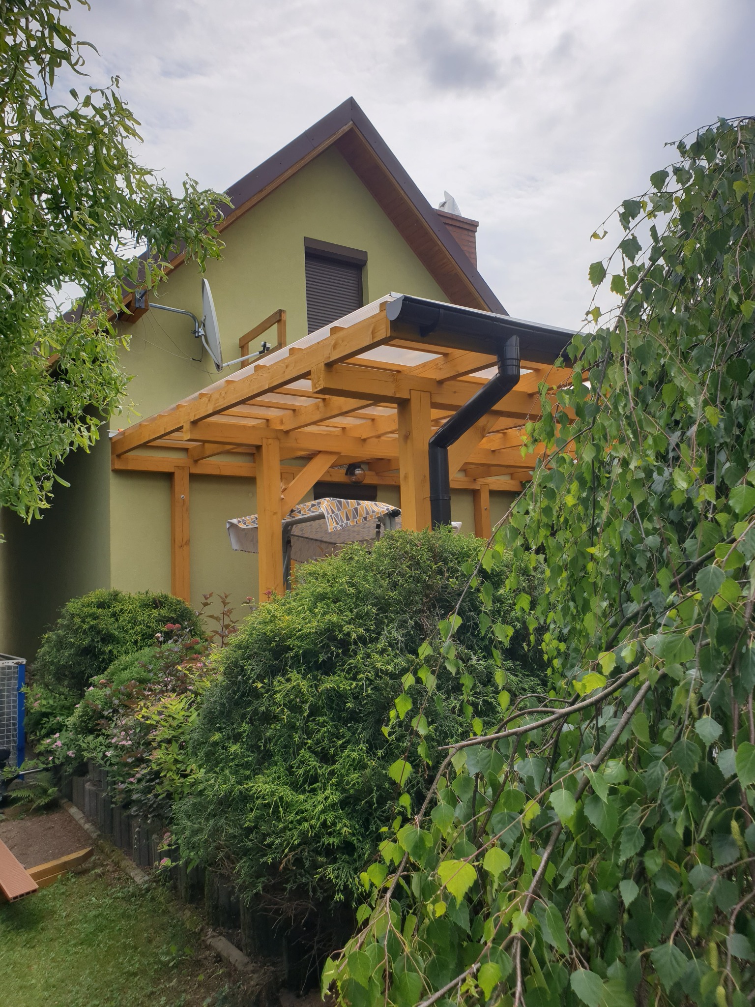 Drewniana konstrukcja z przezroczystym zadaszeniem, prawdopodobnie z poliwęglanu, przylegająca do ściany domu w kolorze oliwkowym, z widoczną rynną, otoczona zielenią.