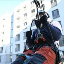 Pracownik wysokościowy w uprzęży, podczas pracy na elewacji nowoczesnego budynku mieszkalnego z balkonami, zabezpieczony linami i kaskiem.