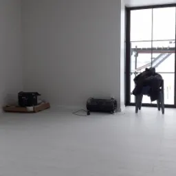 Puste, jasne pomieszczenie z białymi ścianami i podłogą z paneli, widoczny grzejnik elektryczny, kartonowe pudło, krzesło z odzieżą i duże okno z widokiem na budowę.