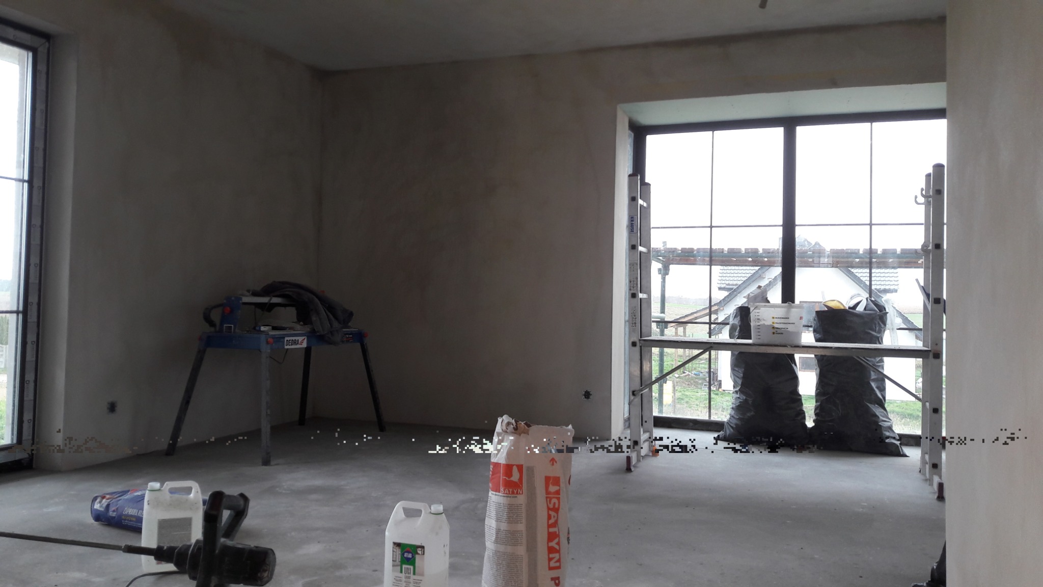 Wnętrze w trakcie prac wykończeniowych: ściany po wstępnym nałożeniu gładzi, widoczne narzędzia, worki z materiałami budowlanymi oraz rusztowanie przy oknie, przez które widać zabudowania.
