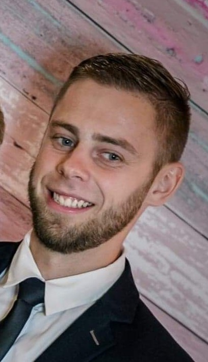 Portret uśmiechniętego mężczyzny z brodą w garniturze na tle ściany z desek w pastelowych kolorach.