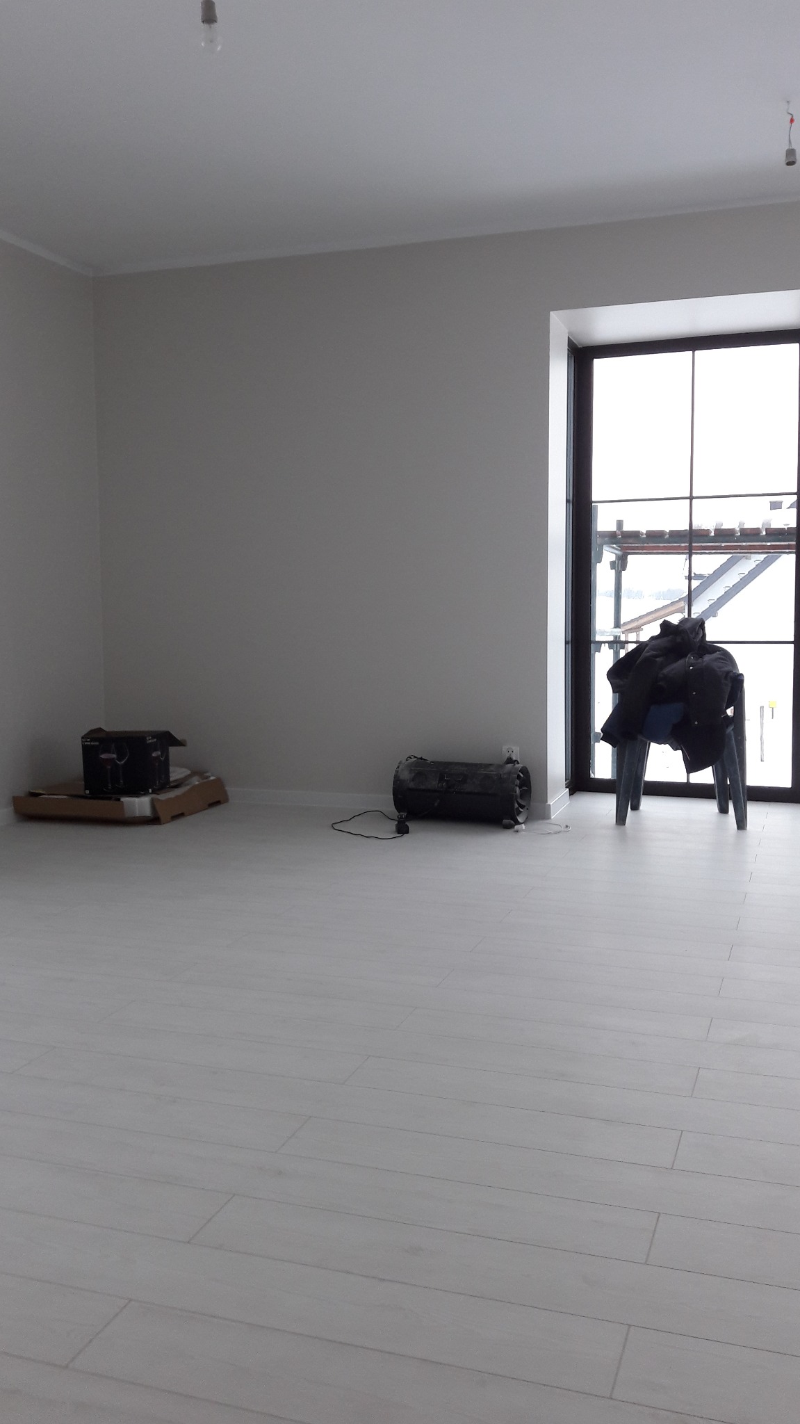 Puste, jasne pomieszczenie z białymi ścianami i podłogą z paneli, widoczny grzejnik elektryczny, kartonowe pudło, krzesło z odzieżą i duże okno z widokiem na budowę.