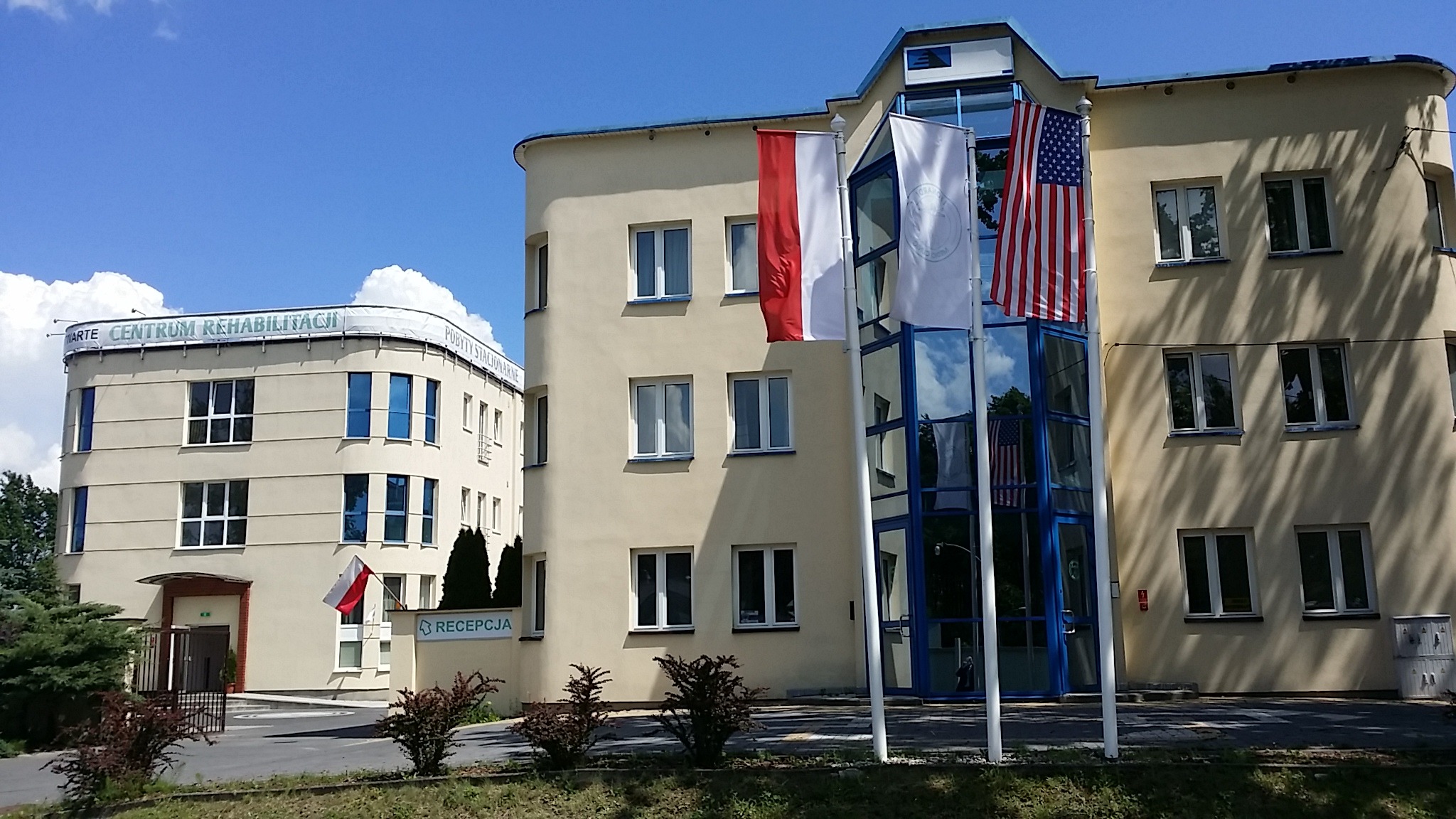 Budynek centrum rehabilitacji z flagami Polski i USA przed wejściem, widoczny napis 'Recepcja'.