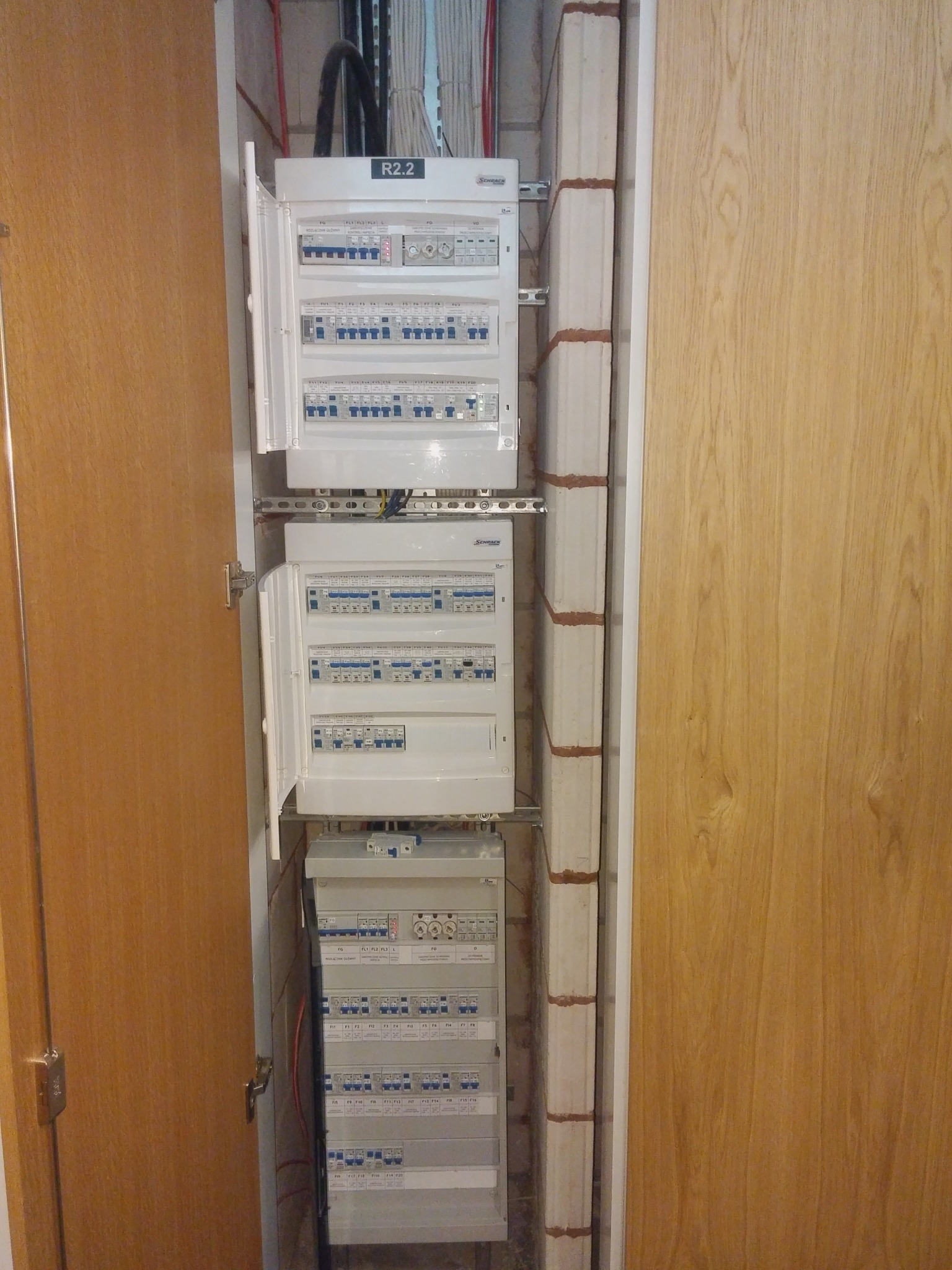 Rozdzielnica elektryczna z otwartymi drzwiczkami, widoczne zabezpieczenia i okablowanie wewnątrz szafy, obok pion instalacyjny z cegieł.