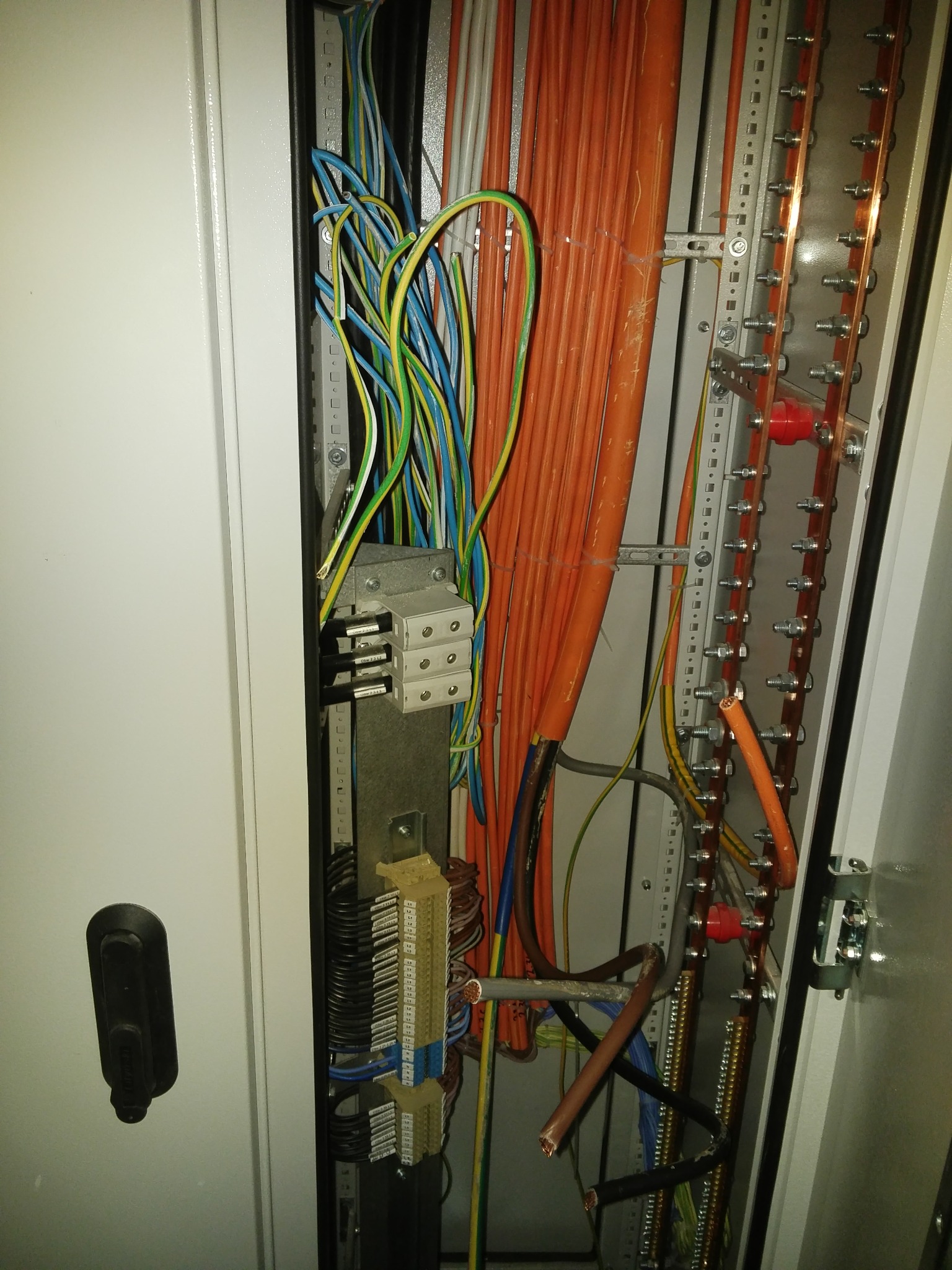 Wnętrze szafy elektrycznej z licznymi przewodami w kolorach pomarańczowym, niebieskim, żółto-zielonym i czarnym, podłączonymi do listew zaciskowych i szyn wyrównawczych z miedzianymi elementami...