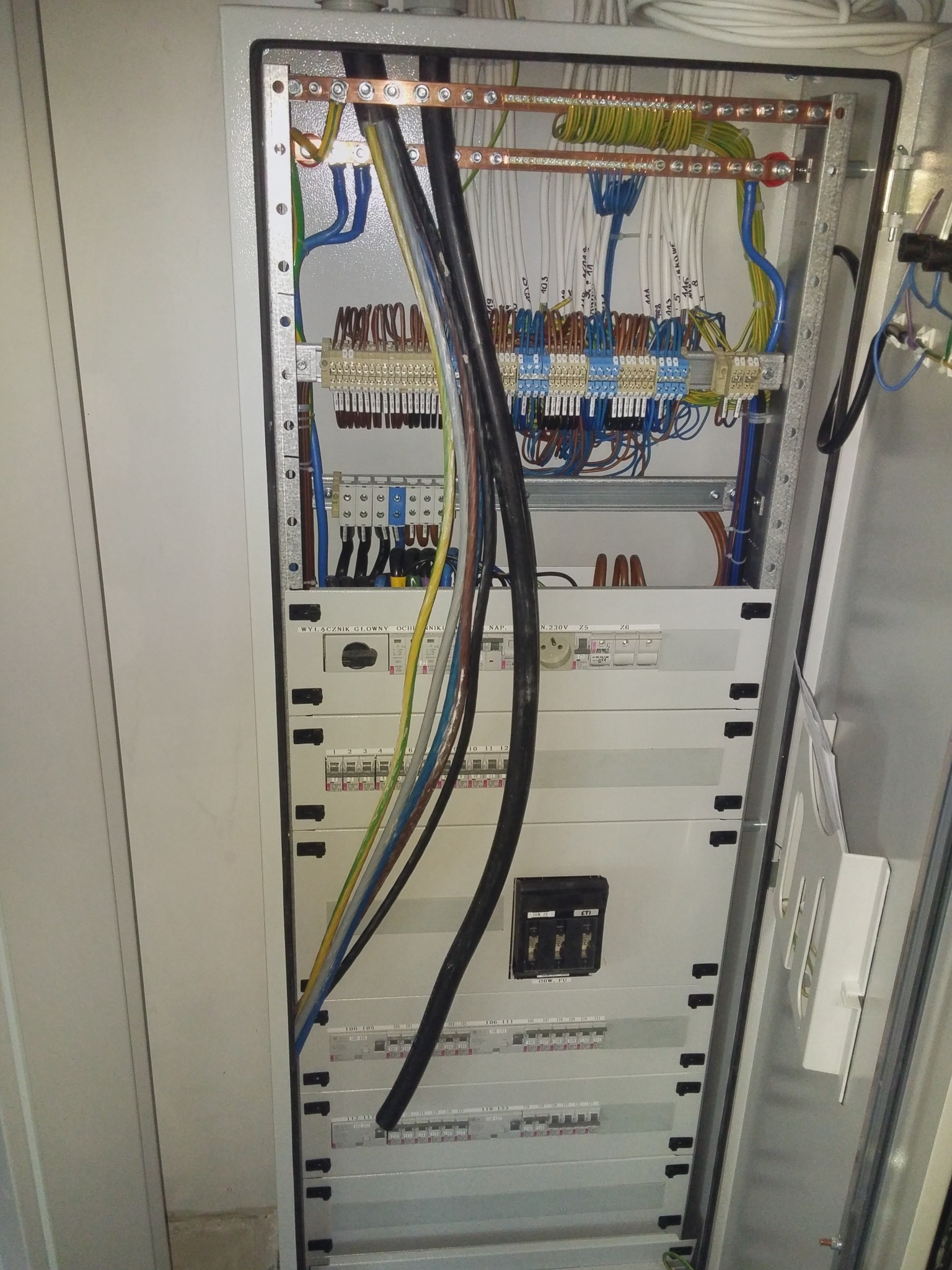 Otwarta szafa elektryczna z widocznymi przewodami, bezpiecznikami i elementami łączeniowymi, okablowanie w kolorach, z opisami na przewodach, widoczne zabezpieczenia i wyłączniki prądowe.