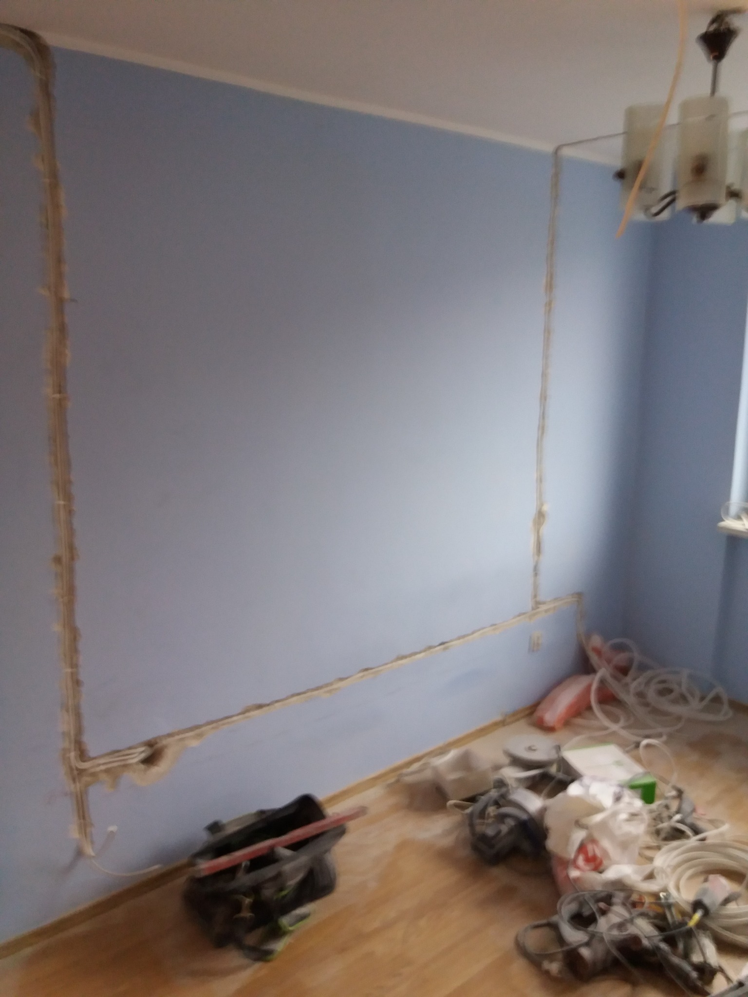 Ściana w pokoju z wyżłobieniami pod kable elektryczne, narzędzia i przewody na podłodze, widoczna lampa sufitowa.