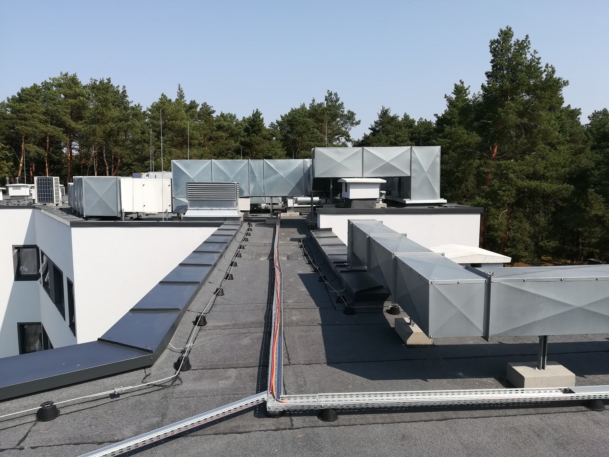 Widok z góry na dach budynku z zainstalowanymi systemami wentylacyjnymi, klimatyzatorami i zabezpieczeniami przeciwprzepięciowymi, na tle drzew.
