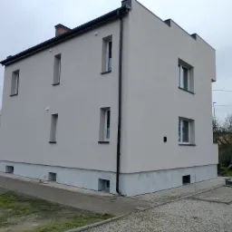 Dwukondygnacyjny budynek mieszkalny z oknami o różnej wysokości, świeżo pomalowaną elewacją w odcieniach beżu i szarości, widok z zewnątrz, pochmurne niebo.