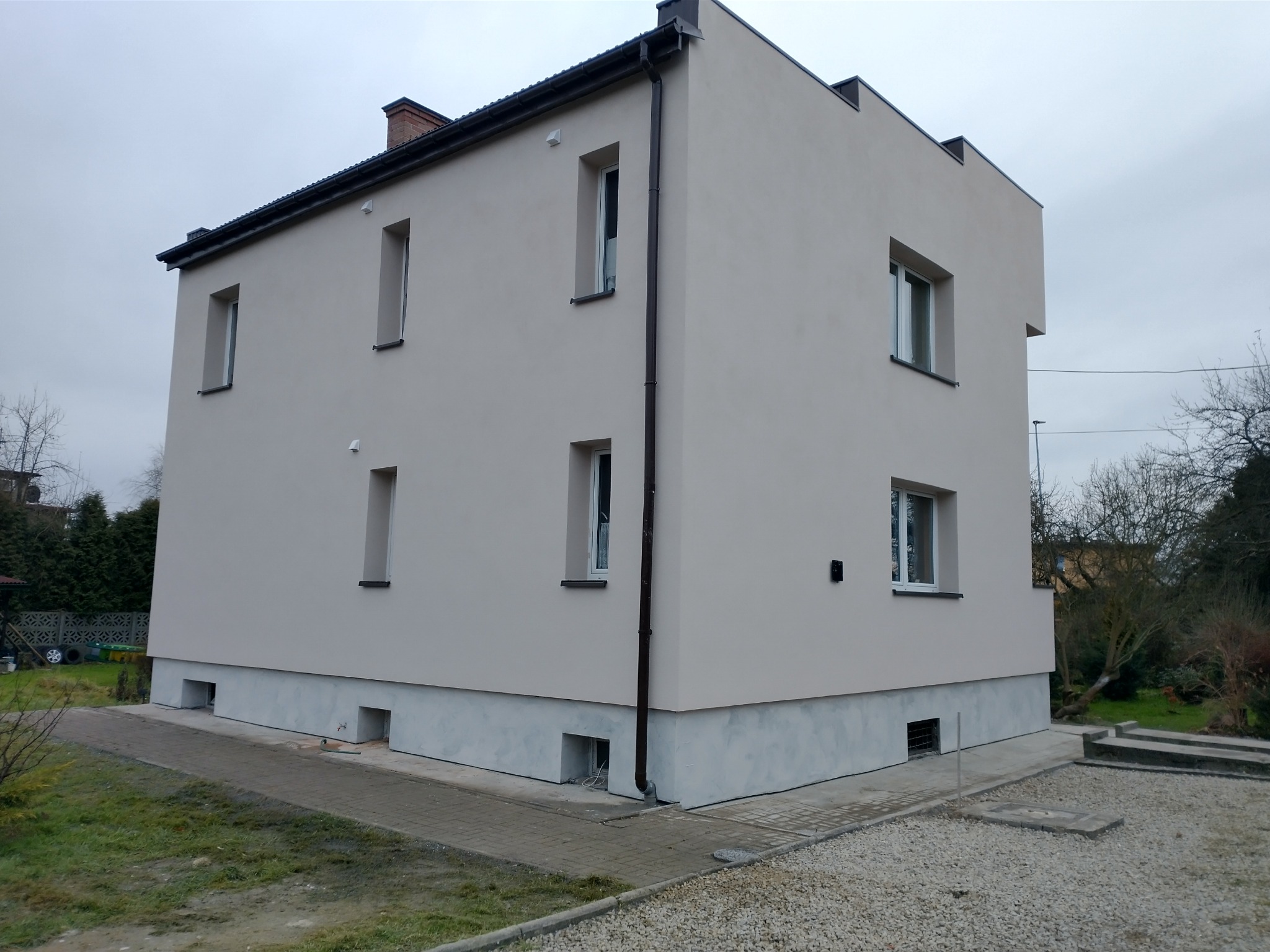 Dwukondygnacyjny budynek mieszkalny z oknami o różnej wysokości, świeżo pomalowaną elewacją w odcieniach beżu i szarości, widok z zewnątrz, pochmurne niebo.