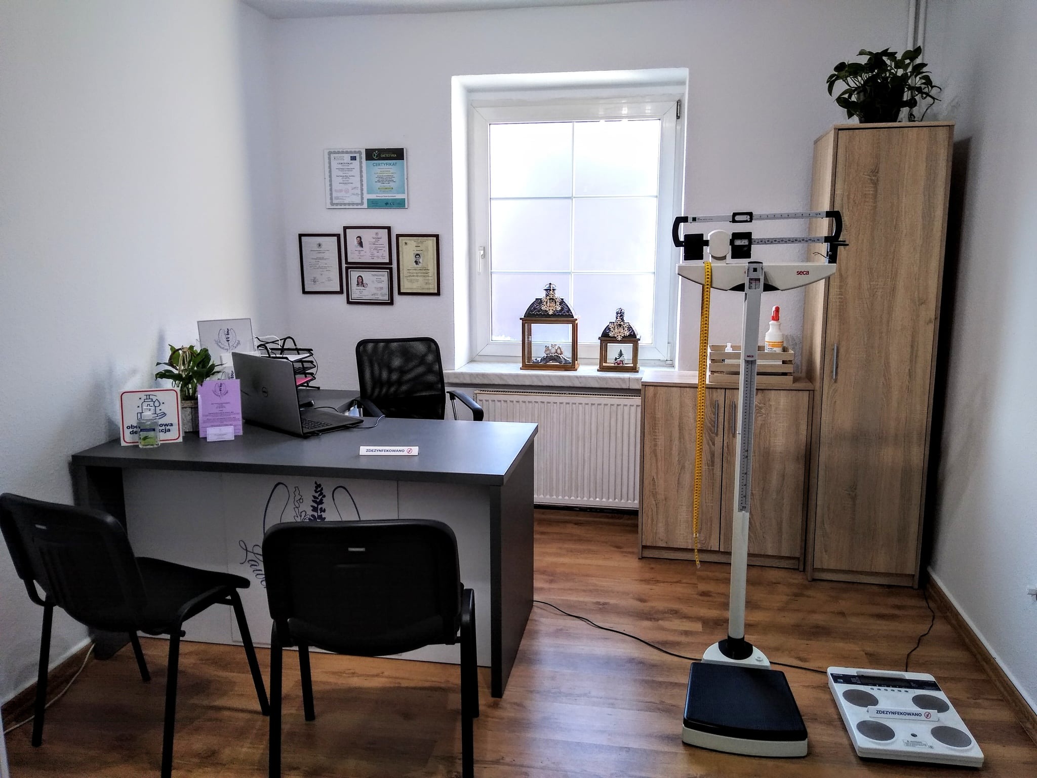 Jasne wnętrze gabinetu z wagą lekarską, biurkiem z laptopem i certyfikatami na ścianie, dekoracyjne lampiony na parapecie okna.