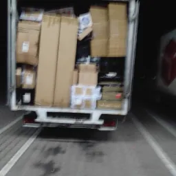 Tył białej furgonetki załadowanej różnej wielkości kartonami, widoczne światła stop i mokra nawierzchnia parkingu.