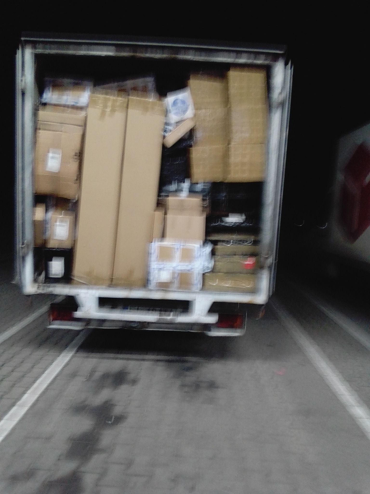 Tył białej furgonetki załadowanej różnej wielkości kartonami, widoczne światła stop i mokra nawierzchnia parkingu.