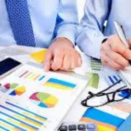 Dwie osoby w błękitnych koszulach analizują kolorowe wykresy i notują dane, używając długopisów na białym biurku z kalkulatorami i okularami.