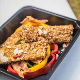 Kwadratowe czarne pudełko z trzema batonami zbożowymi posypanymi płatkami migdałów, ułożonymi na grillowanej papryce: czerwonej, żółtej i zielonej, oraz boczku.